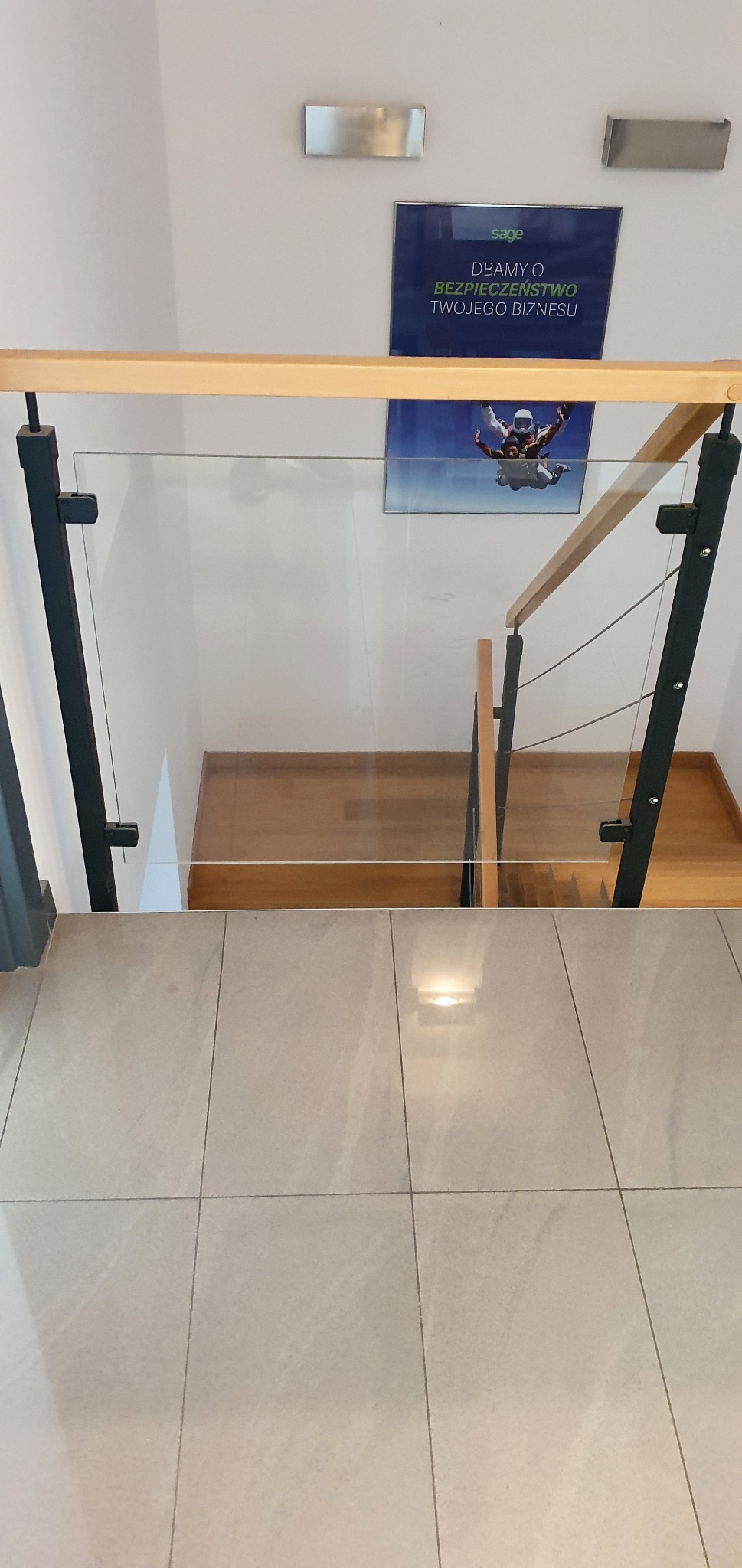 Widok z góry na schody drewniane z balustradą ze szkła i elementami metalowymi, na ścianie plakat z motywem skoków spadochronowych.