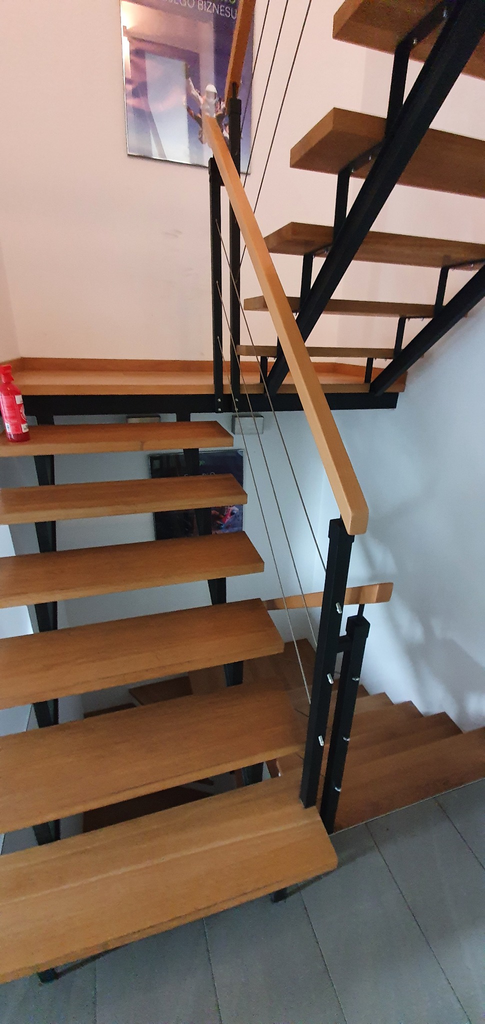 Nowoczesne schody z drewnianymi stopniami i czarną metalową konstrukcją, z jasną drewnianą poręczą i stalowymi linkami zabezpieczającymi, częściowo widoczny plakat na ścianie.