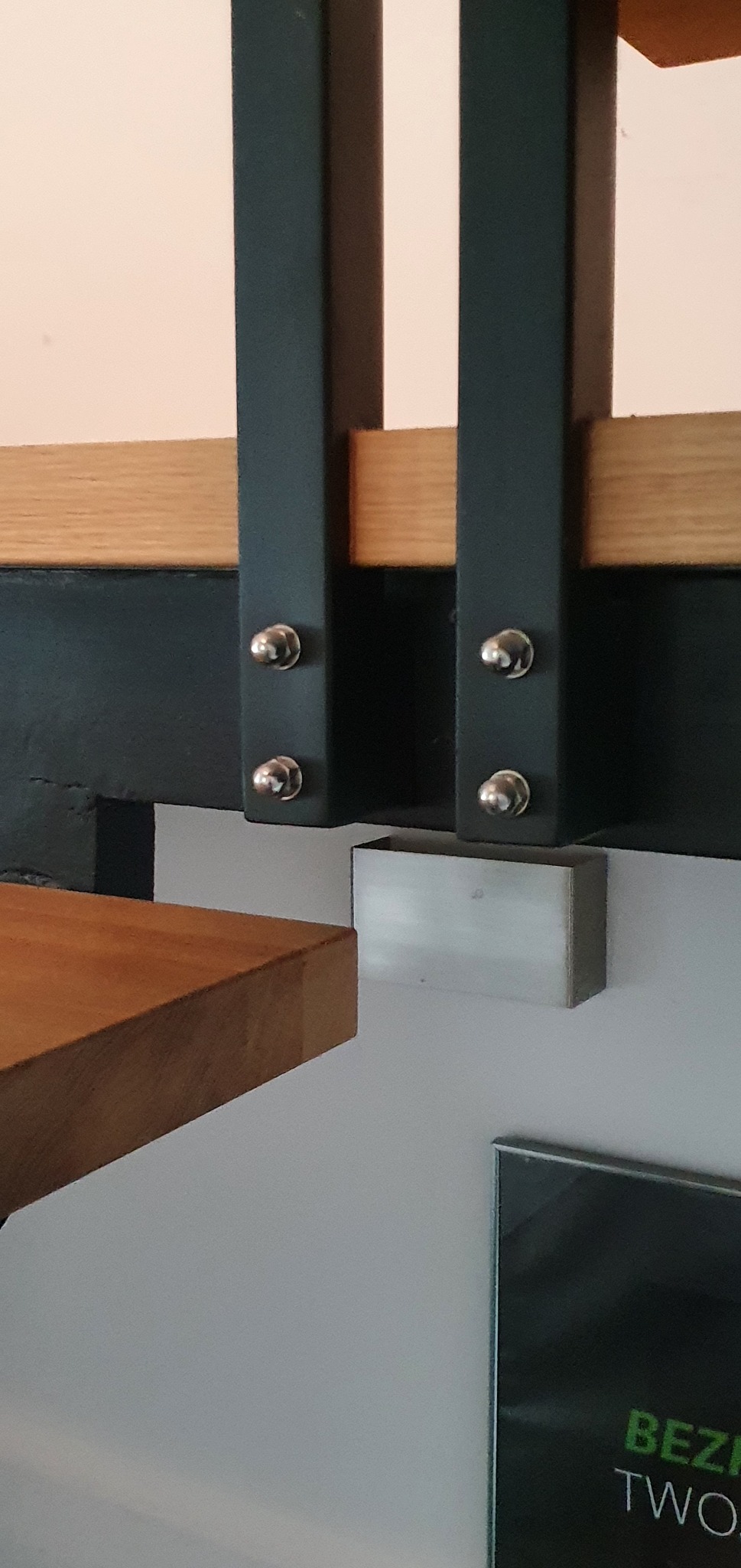 Fragment konstrukcji schodów z drewnianymi stopniami, czarnymi metalowymi wspornikami i srebrnymi śrubami mocującymi, zbliżenie na połączenie elementów.