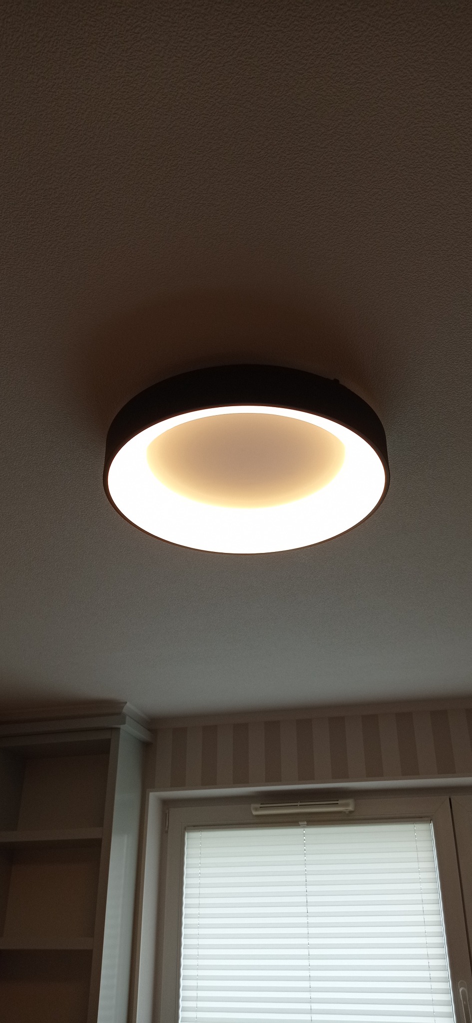 Zamontowana okrągła lampa sufitowa LED z czarną obudową, emitująca ciepłe białe światło, widoczna na tle sufitu i fragmentu pokoju z tapetą w paski oraz oknem z żaluzjami.