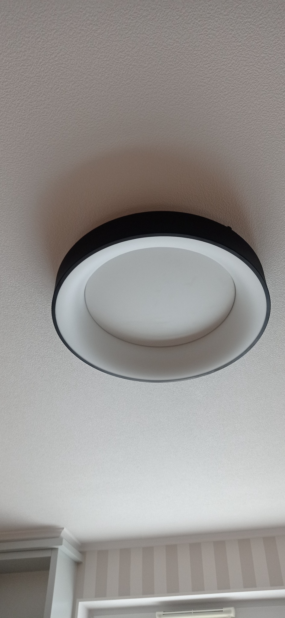 Okrągła lampa sufitowa LED z czarną obudową i białym kloszem, zamontowana na jasnym suficie, widoczny fragment ściany w paski.