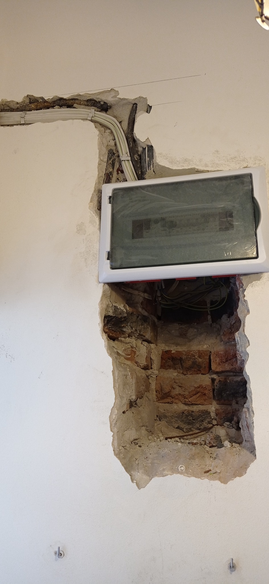 Niefortunne połączenie: nowa skrzynka elektryczna z folią ochronną, osadzona w surowej dziurze w ścianie z odsłoniętą cegłą, widoczne kable elektryczne u góry.
