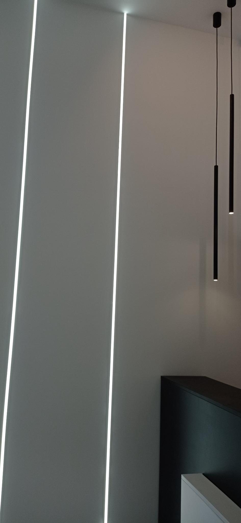 Nowoczesne oświetlenie wnętrza: pionowe, wpuszczane listwy LED w ścianie i wiszące lampy tubowe z czarnymi przewodami.