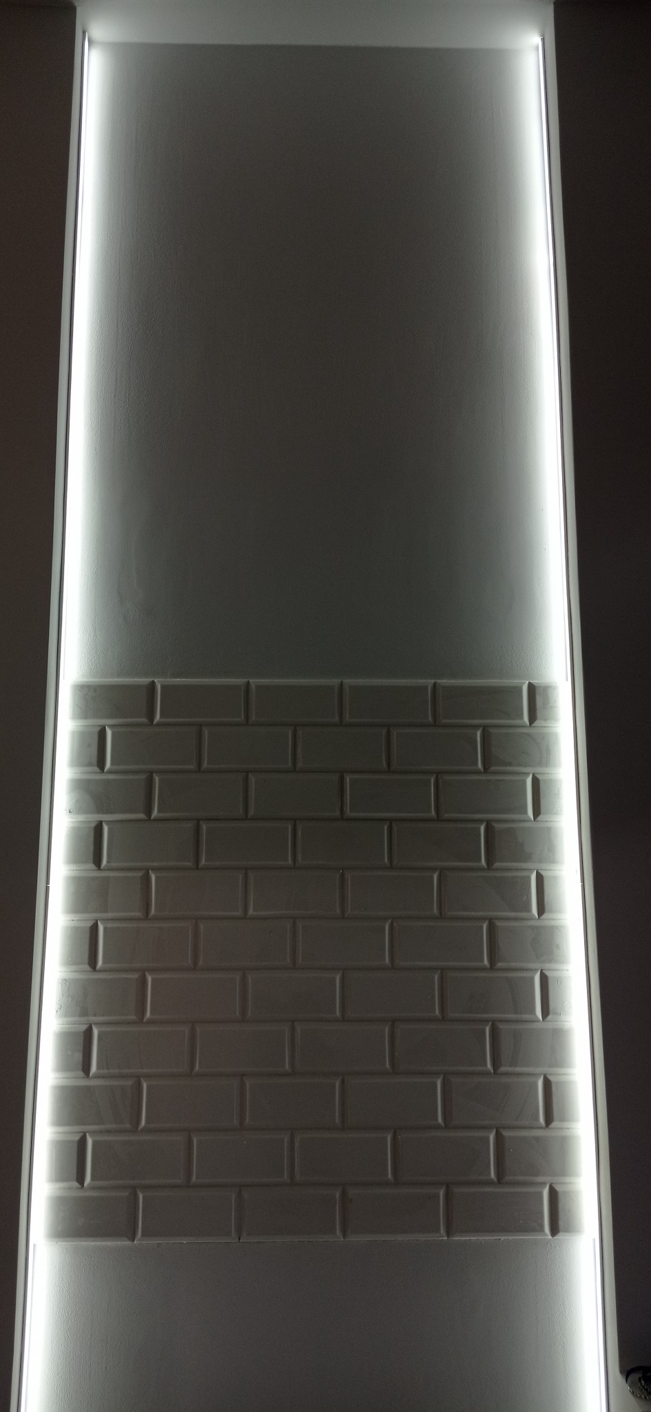 Ściana z białych, prostokątnych płytek, oświetlona pionowymi paskami LED po bokach, tworzącymi jasną ramę.