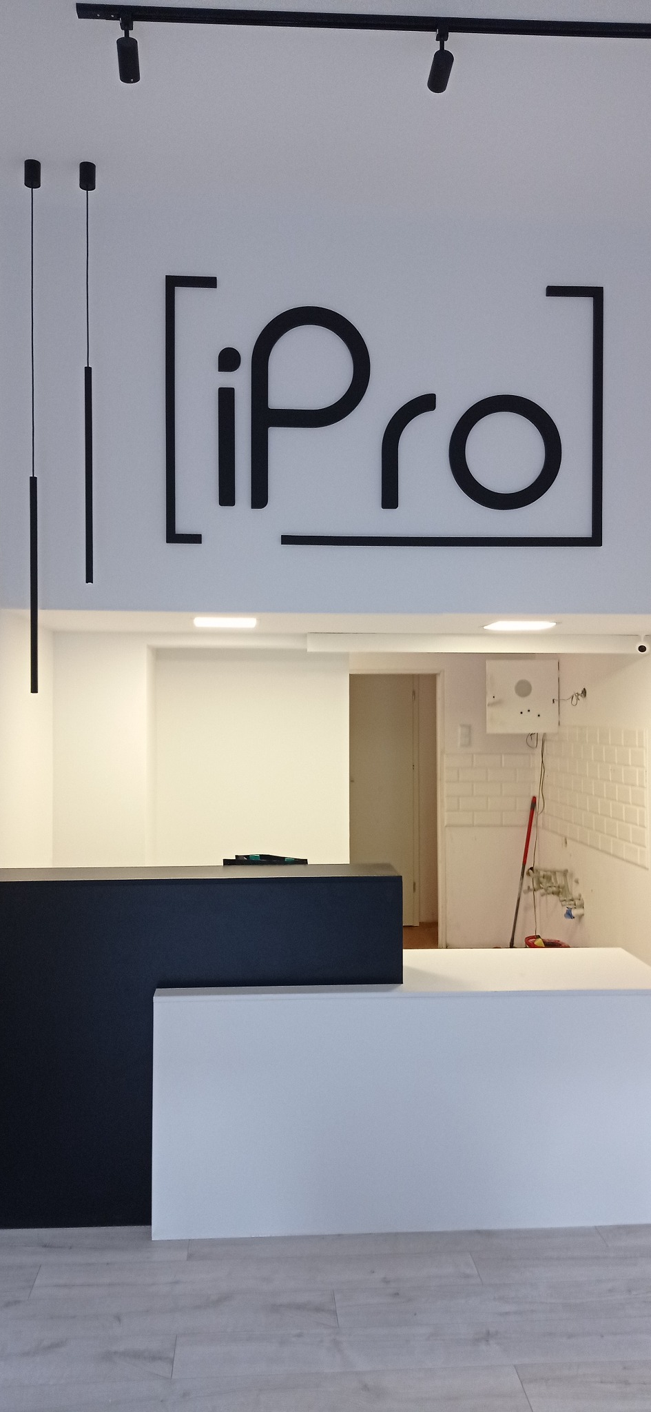 Recepcja z logo firmy 'iPro' pod nowoczesnym oświetleniem punktowym i wiszącymi lampami cylindrycznymi; w tle ściana z białej cegły i instalacja elektryczna w trakcie prac.