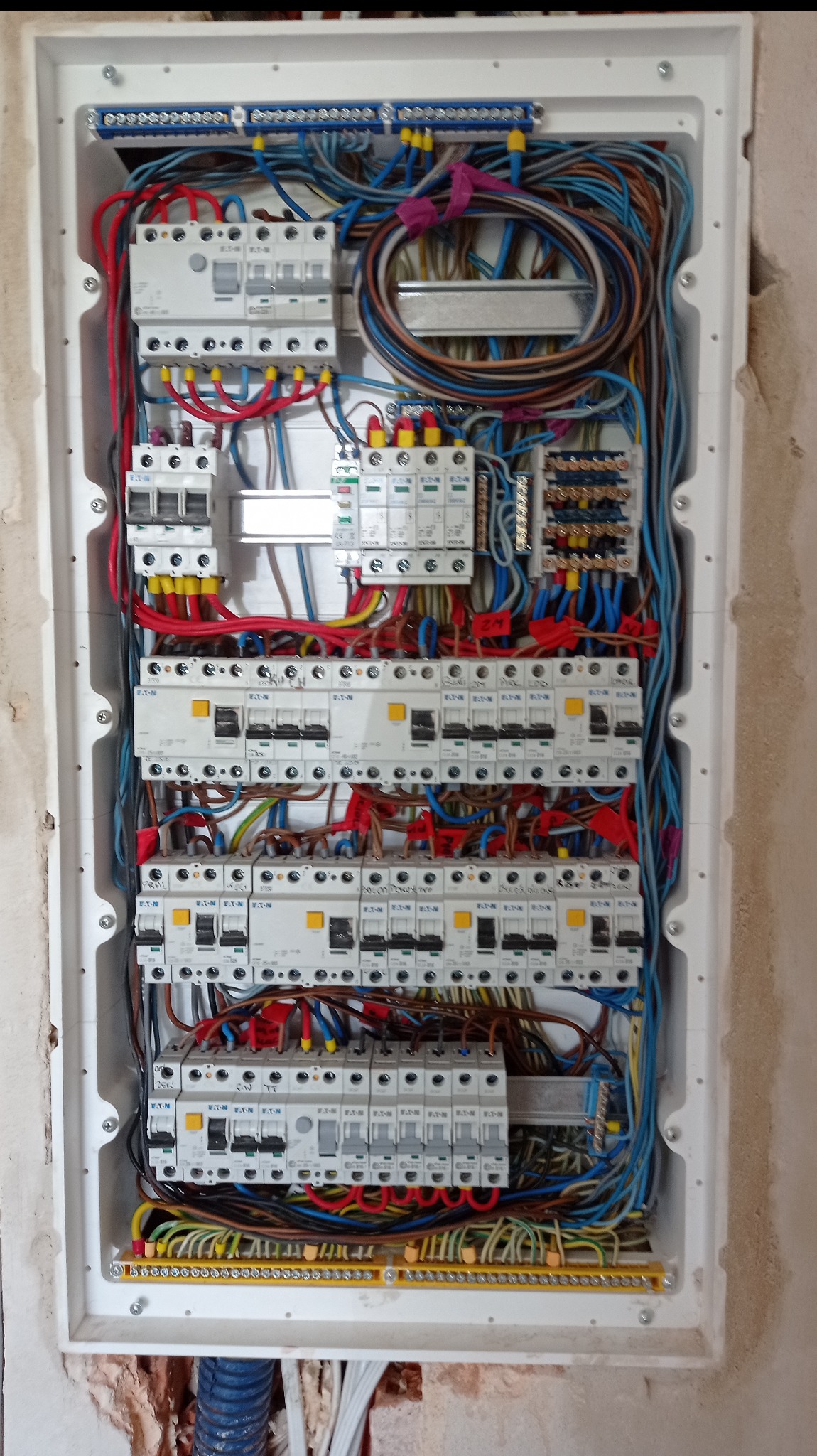 Rozdzielnica elektryczna w białej obudowie, widoczne liczne bezpieczniki, przewody w różnych kolorach (niebieski, czerwony, brązowy, żółto-zielony) oraz listwy zaciskowe. Instalacja w trakcie...