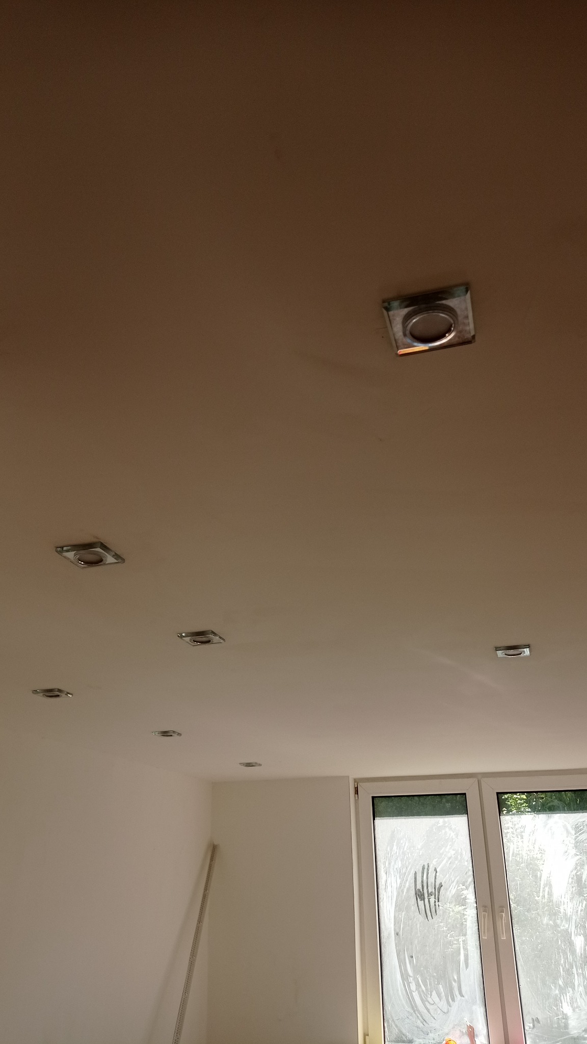 Biały sufit z zamontowanymi, kwadratowymi oprawami oświetleniowymi halogenowymi, widok z dołu, okno w tle.