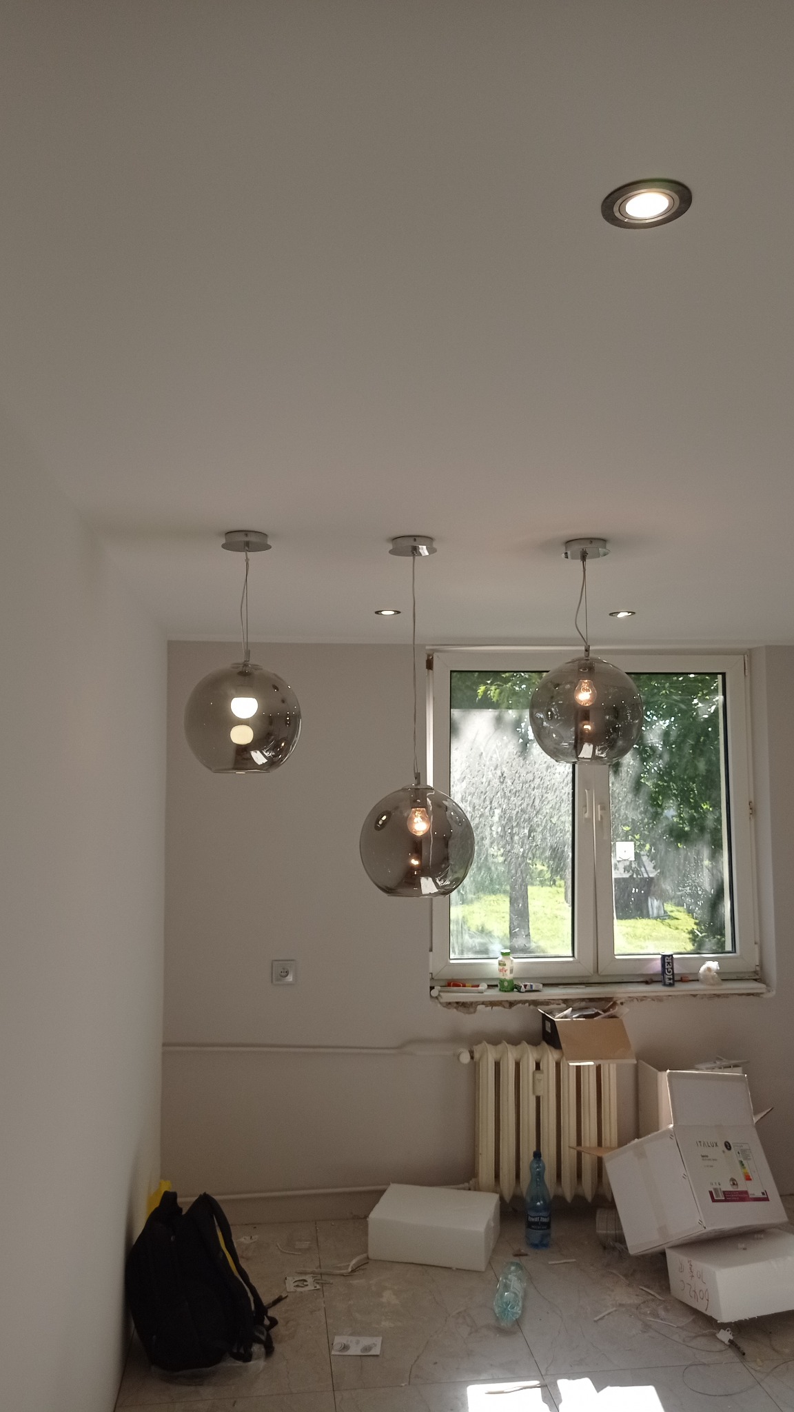 Trzy szklane lampy wiszące w kształcie kuli, zamontowane w pomieszczeniu z widokiem na zieleń za oknem; widoczne kartony po montażu i narzędzia na podłodze.