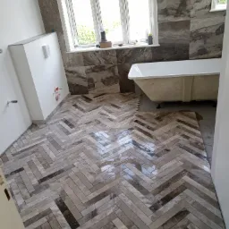 Ułożona w jodełkę, połyskująca, marmurowa podłoga w łazience, z widoczną wanną i fragmentem okna. Częściowo zakończone prace glazurnicze.