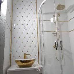 Biała łazienka z kabiną prysznicową i umywalką w kolorze złota. Ściany wyłożone białymi płytkami z dekoracyjnymi, złotymi elementami.