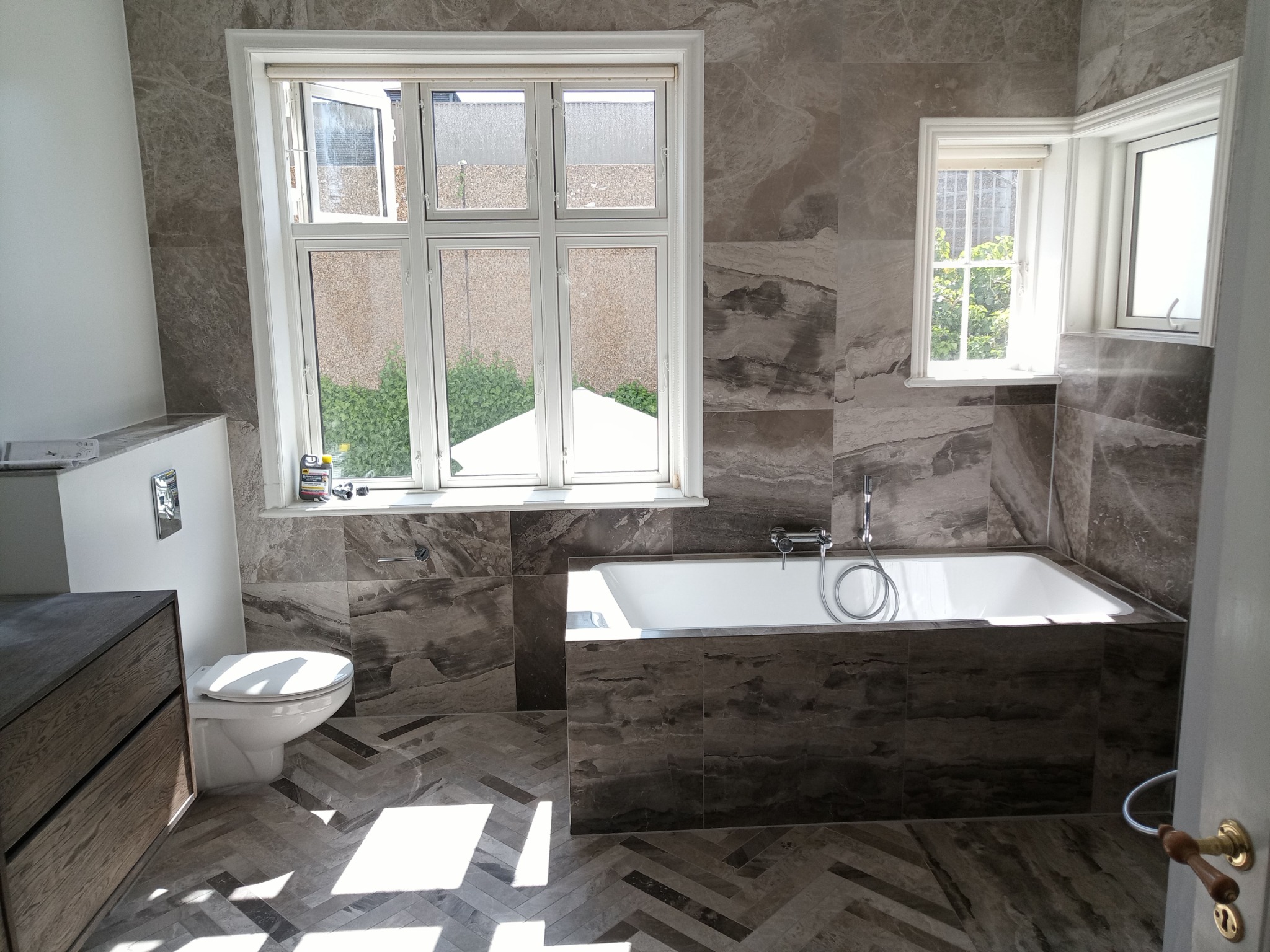 Elegancka łazienka z marmurowymi płytkami, wzorem w jodełkę na podłodze i oknami wpuszczającymi naturalne światło. Wanna obudowana płytkami, umywalka z szafką.