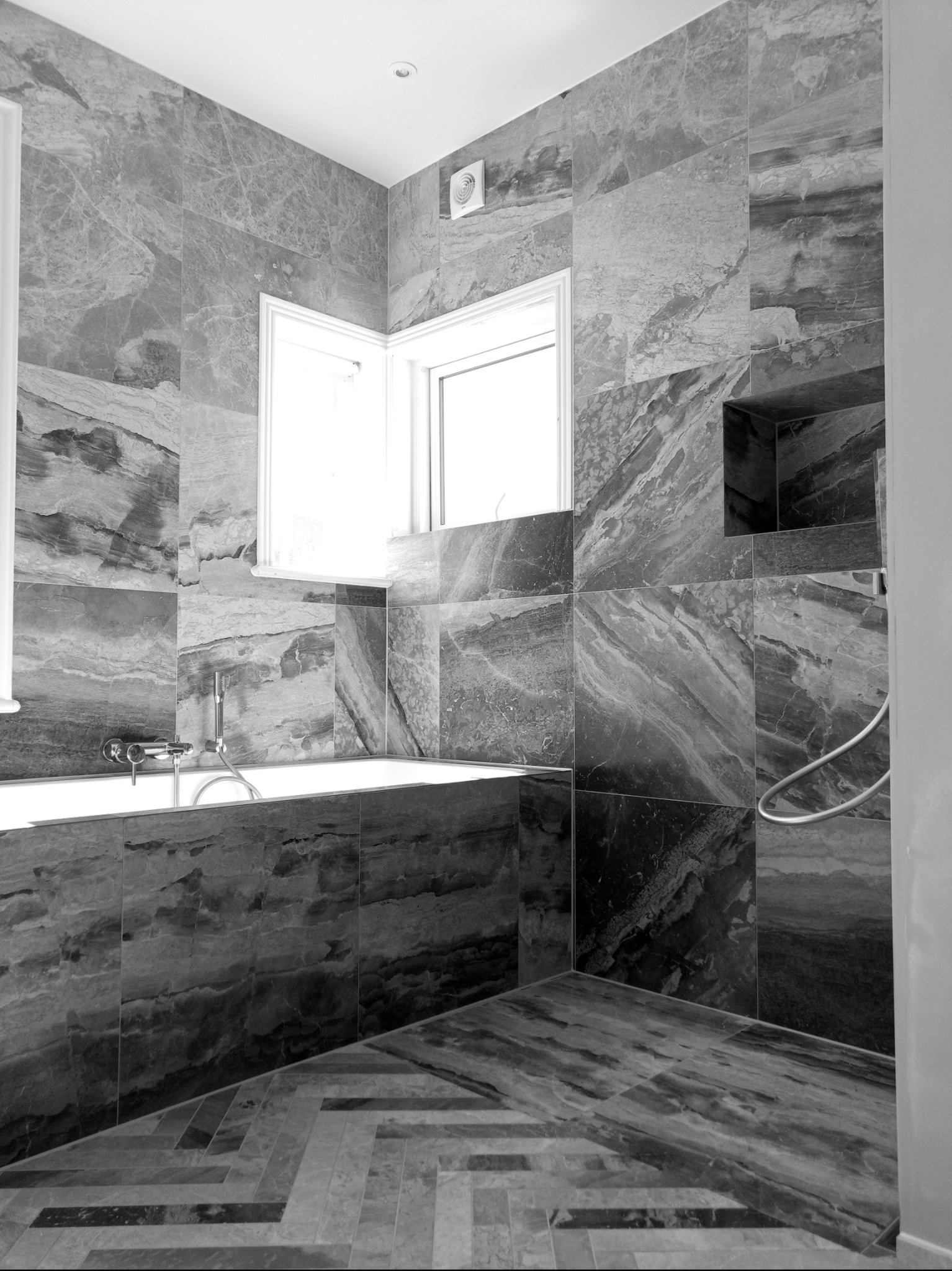 Elegancka łazienka w odcieniach szarości z marmurowymi płytkami, widoczna wanna, okno i mozaika na podłodze. Minimalistyczny design i nowoczesne wykończenie.