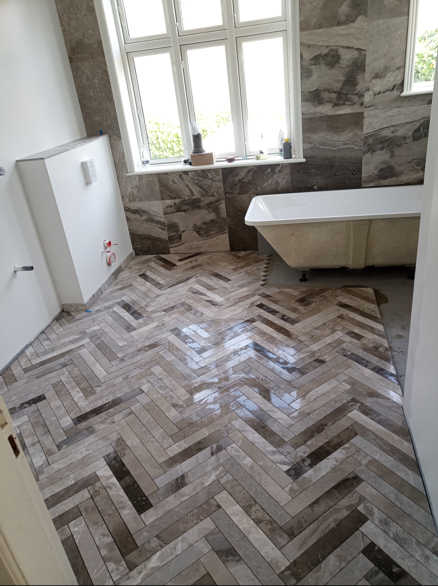 Ułożona w jodełkę, połyskująca, marmurowa podłoga w łazience, z widoczną wanną i fragmentem okna. Częściowo zakończone prace glazurnicze.