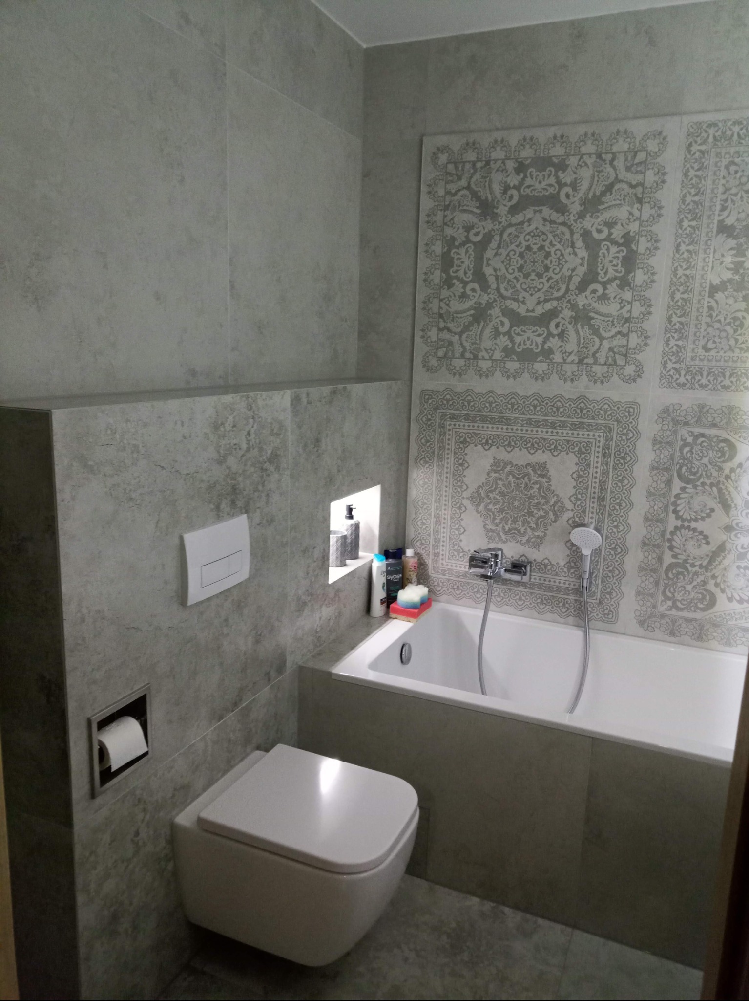 Wykończona łazienka w szarościach: nowoczesna toaleta podtynkowa, wanna z baterią i deszczownicą, dekoracyjne płytki nad wanną, wnęka z kosmetykami. Minimalistyczny design.