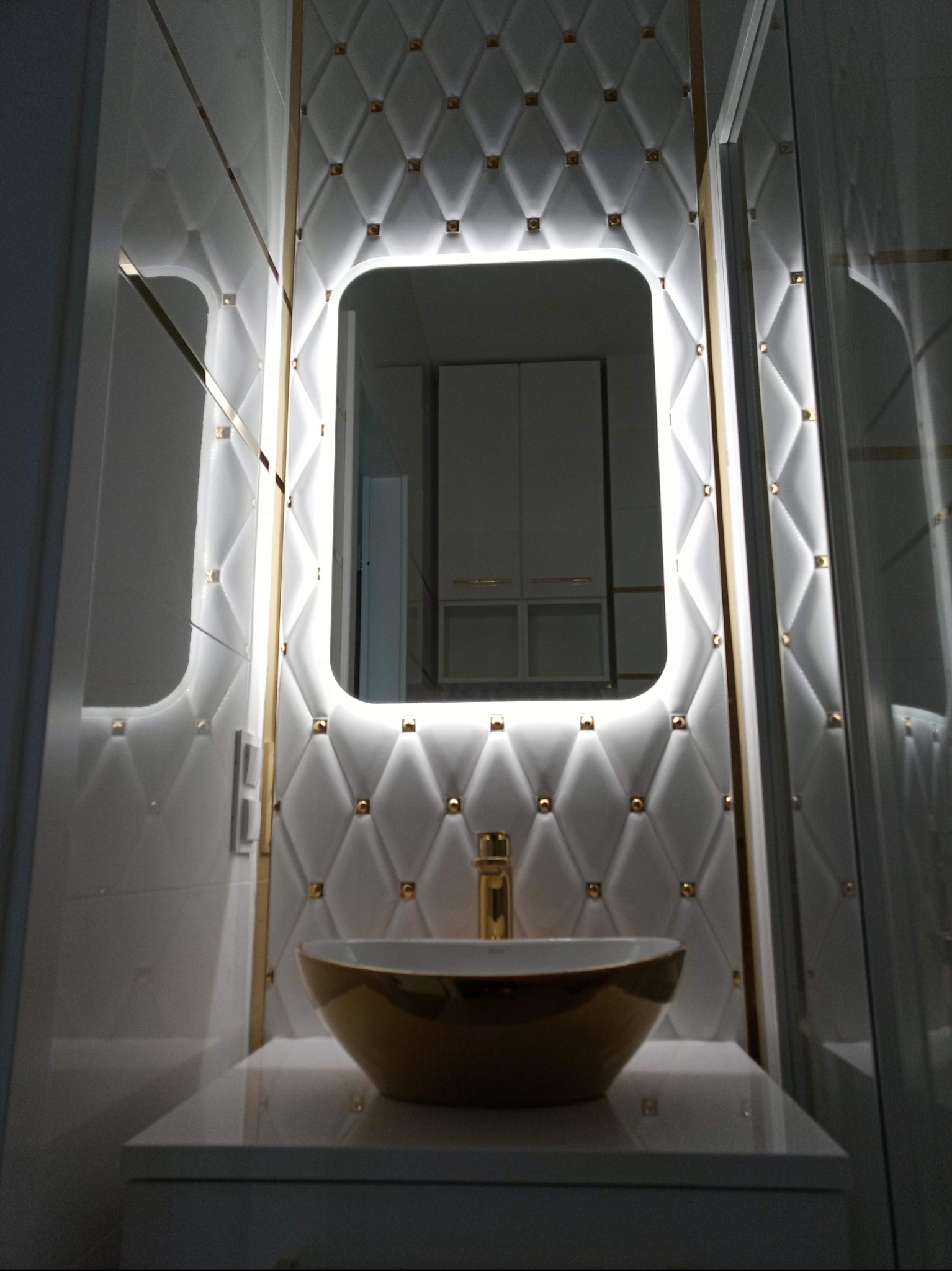 Elegancka łazienka: złota umywalka na białym blacie, lustro z podświetleniem LED i pikowana ściana w romby z złotymi akcentami.