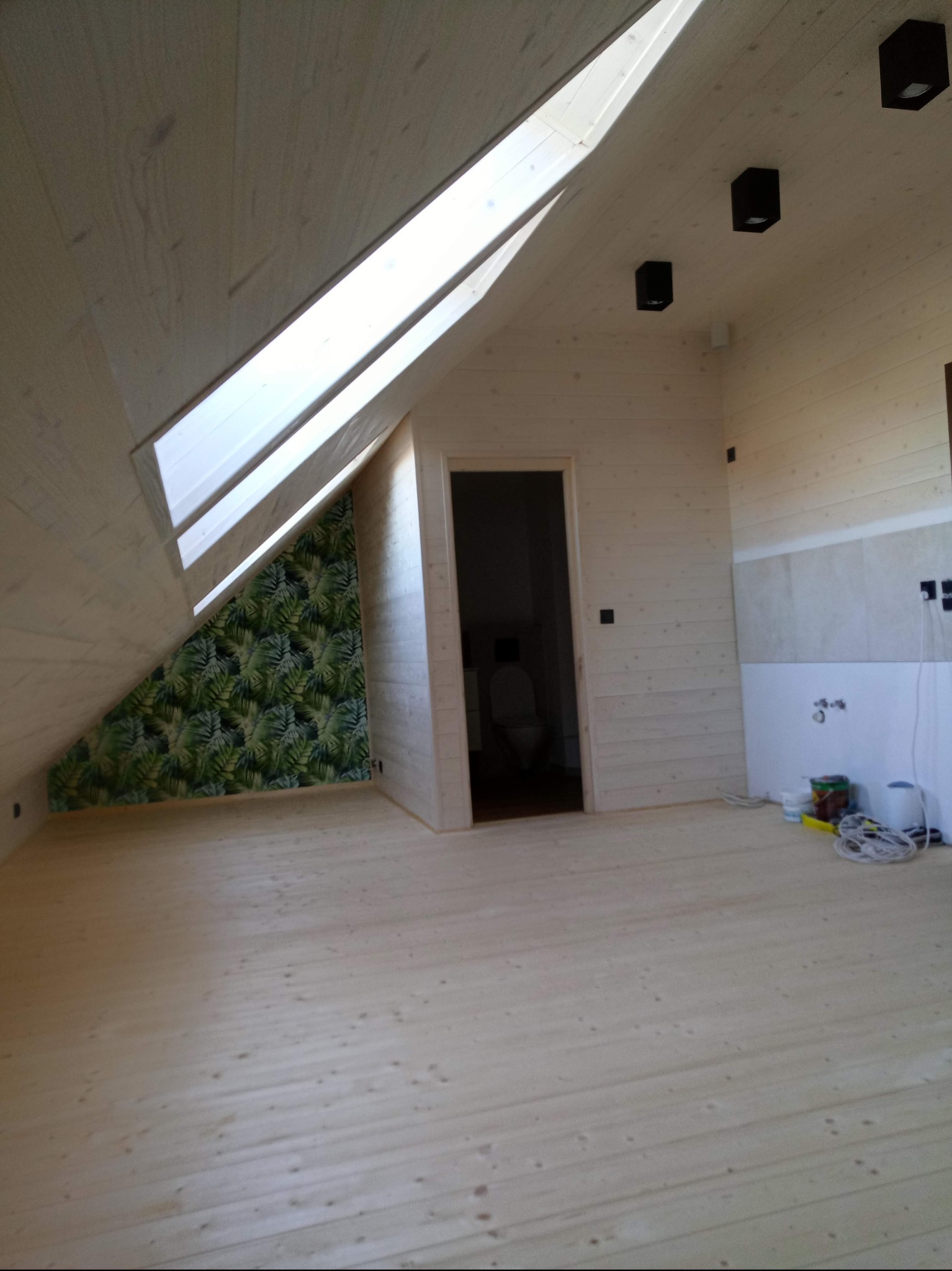 Poddasze po remoncie: drewniane panele na podłodze i ścianach, okno dachowe, ściana z tapetą w liście. Częściowo wykończona łazienka w tle.