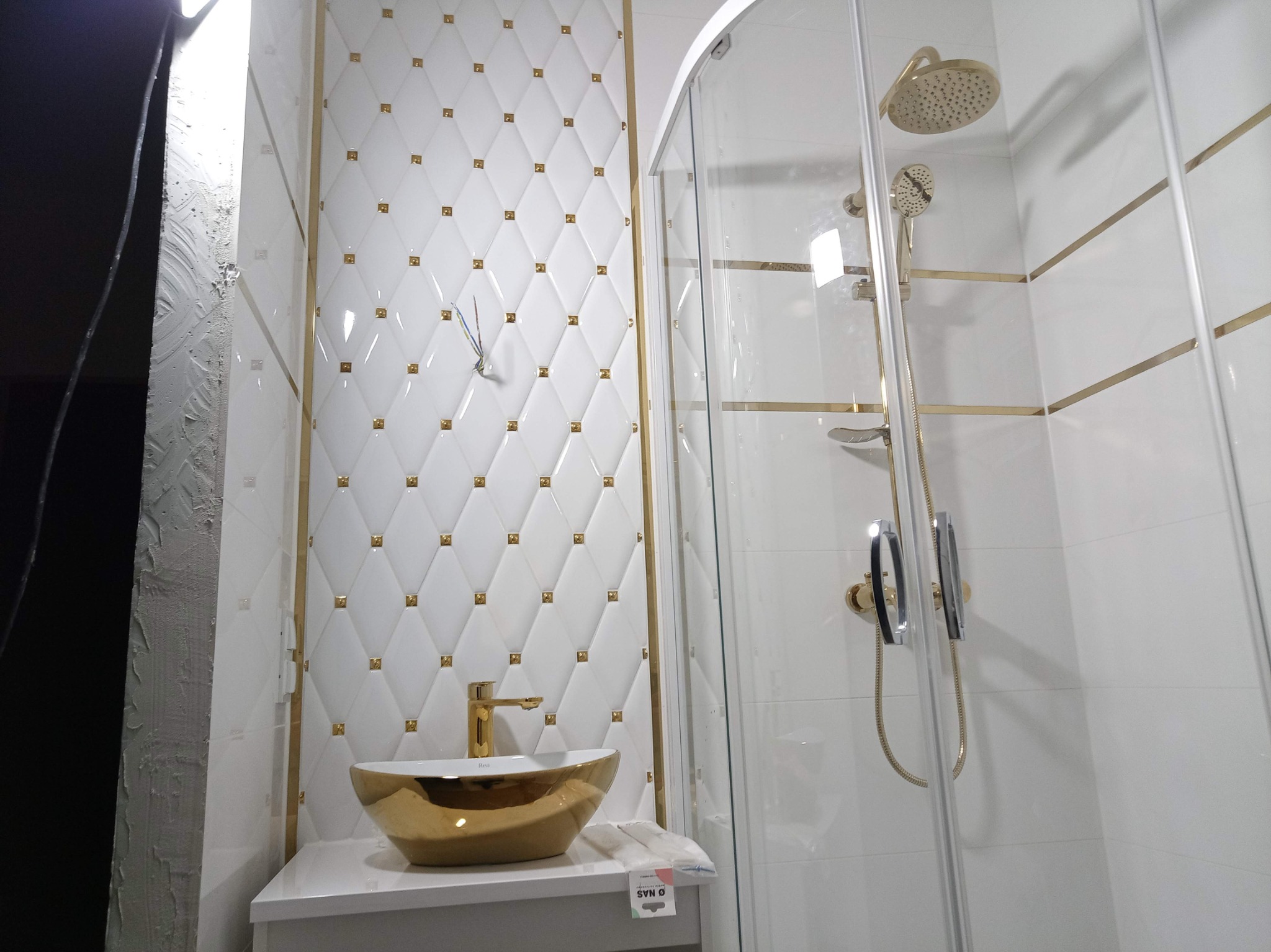 Biała łazienka z kabiną prysznicową i umywalką w kolorze złota. Ściany wyłożone białymi płytkami z dekoracyjnymi, złotymi elementami.