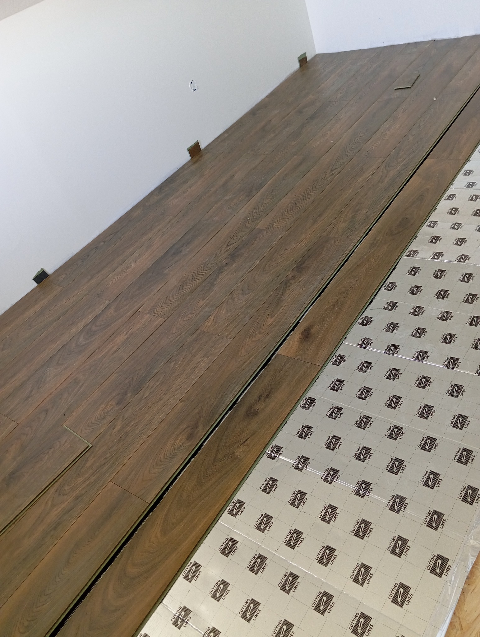 Układanie paneli podłogowych w pomieszczeniu, widoczna podkładka izolacyjna z nadrukiem 'CUTTING LINES'. Częściowo ułożona podłoga z brązowych paneli.