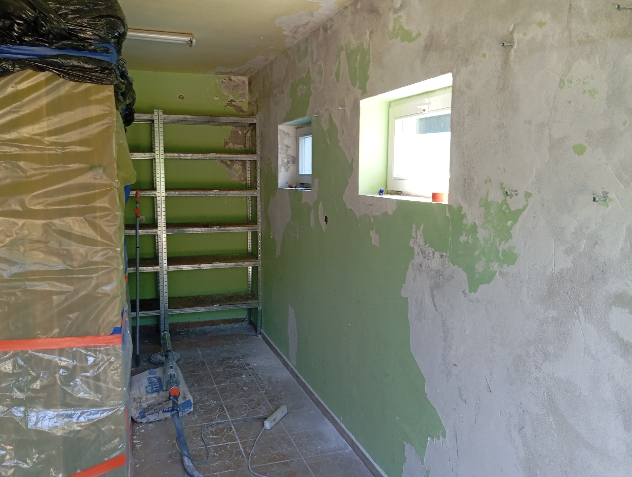 Pokój w trakcie remontu: ściany z resztkami farby, częściowo zagruntowane, metalowy regał, okna. Przygotowania do malowania, zabezpieczone elementy.