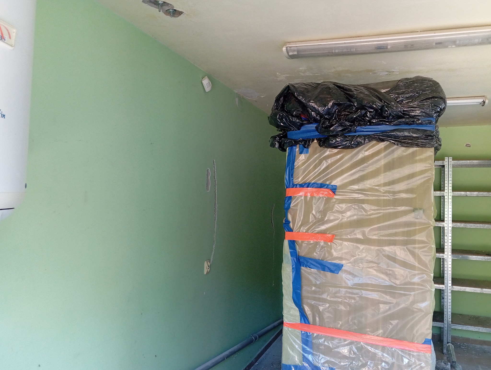 Przygotowanie do remontu: zabezpieczone folią i taśmą drzwi w pomieszczeniu z zieloną ścianą, obok regał metalowy. Widoczne ślady po instalacji na suficie.