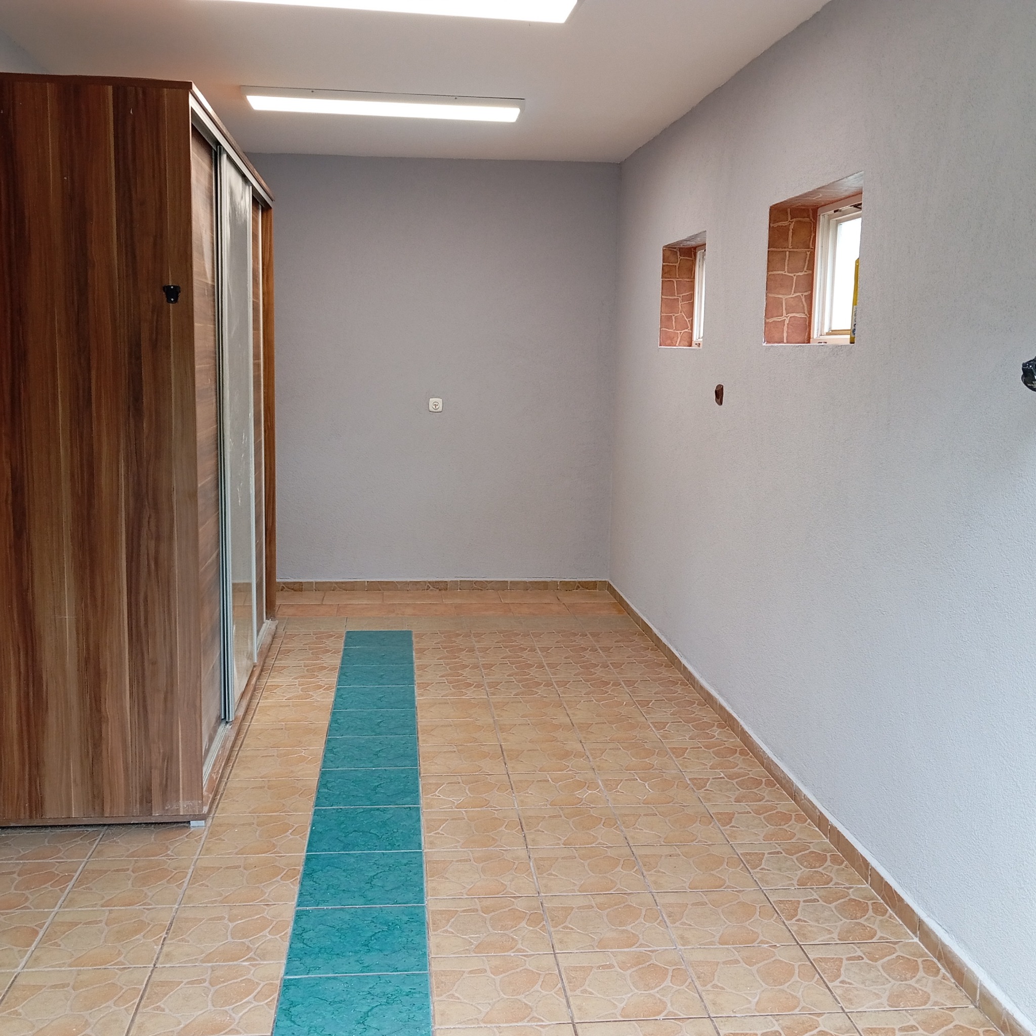 Pokój po remoncie: szara ściana, brązowe płytki z turkusowym pasem, drewniana szafa przesuwna. Dwa okna z obramowaniem imitującym cegłę.