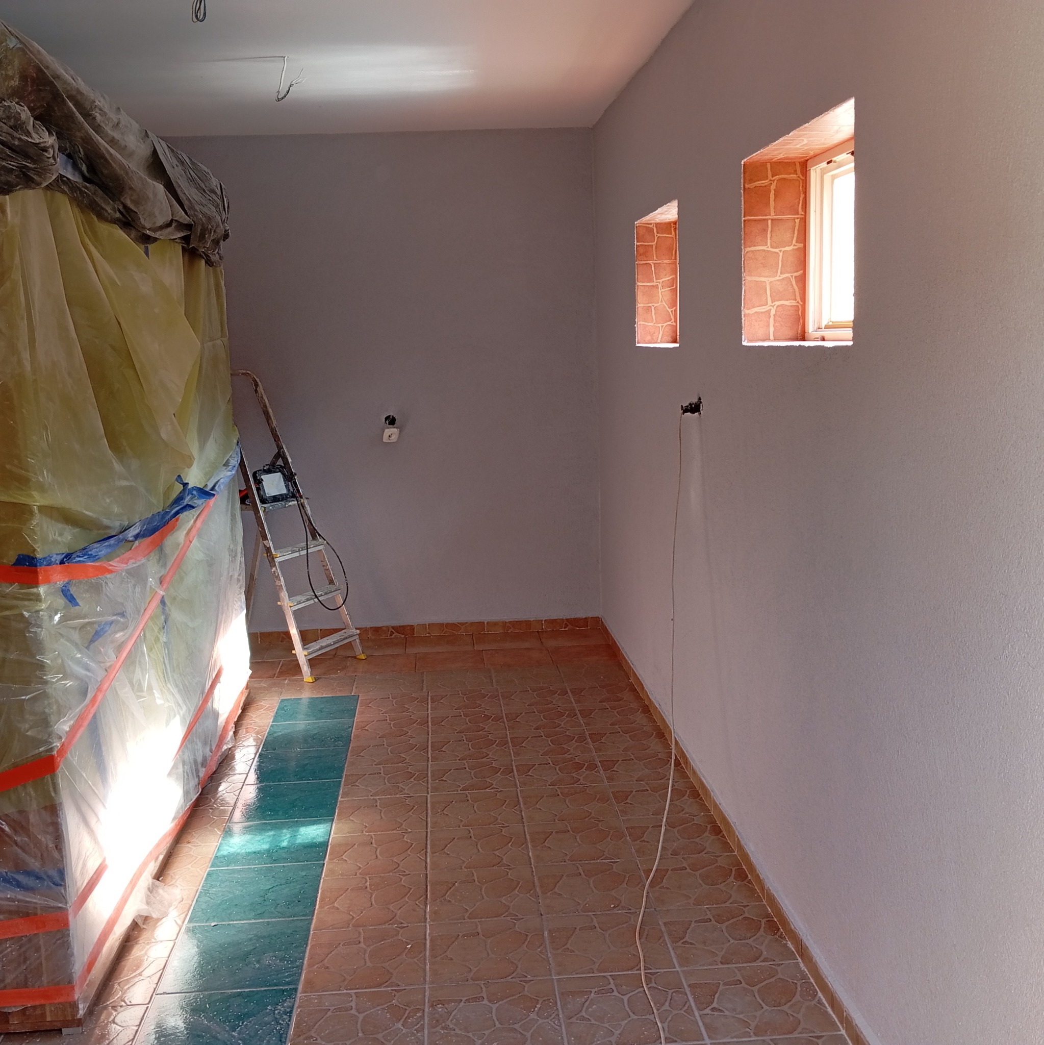 Wnętrze po malowaniu ścian na szaro, zabezpieczone meble folią, widoczne małe okna z ceglanym obramowaniem i drabina. Podłoga wyłożona brązowymi płytkami.