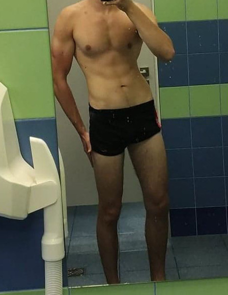 Mężczyzna pozujący w czarnych spodenkach, prezentujący umięśnioną sylwetkę w łazience z niebiesko-zielonymi kafelkami, widoczne odbicie w lustrze.