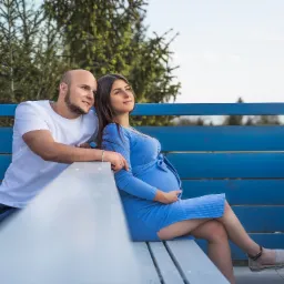 Przyszła mama w błękitnej sukience siedzi na niebieskiej ławce z partnerem, oboje patrzą w dal, drzewa w tle.