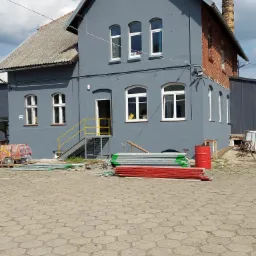 Odnowiona elewacja budynku w odcieniach szarości z ceglanym kominem, widoczne rusztowania i materiały budowlane na kostce brukowej przed budynkiem.