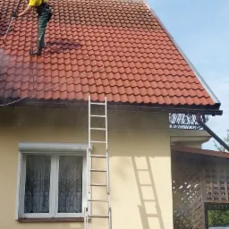 Pracownik w kasku i kombinezonie roboczym czyści dach z czerwonej dachówki za pomocą myjki ciśnieniowej, stojąc na jego krawędzi; widoczna drabina oparta o ścianę budynku.