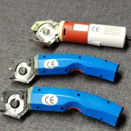 Trzy elektryczne noże tarczowe do cięcia tkanin, jeden z czerwono-białą obudową, dwa z niebieską, leżące na szarej tkaninie.