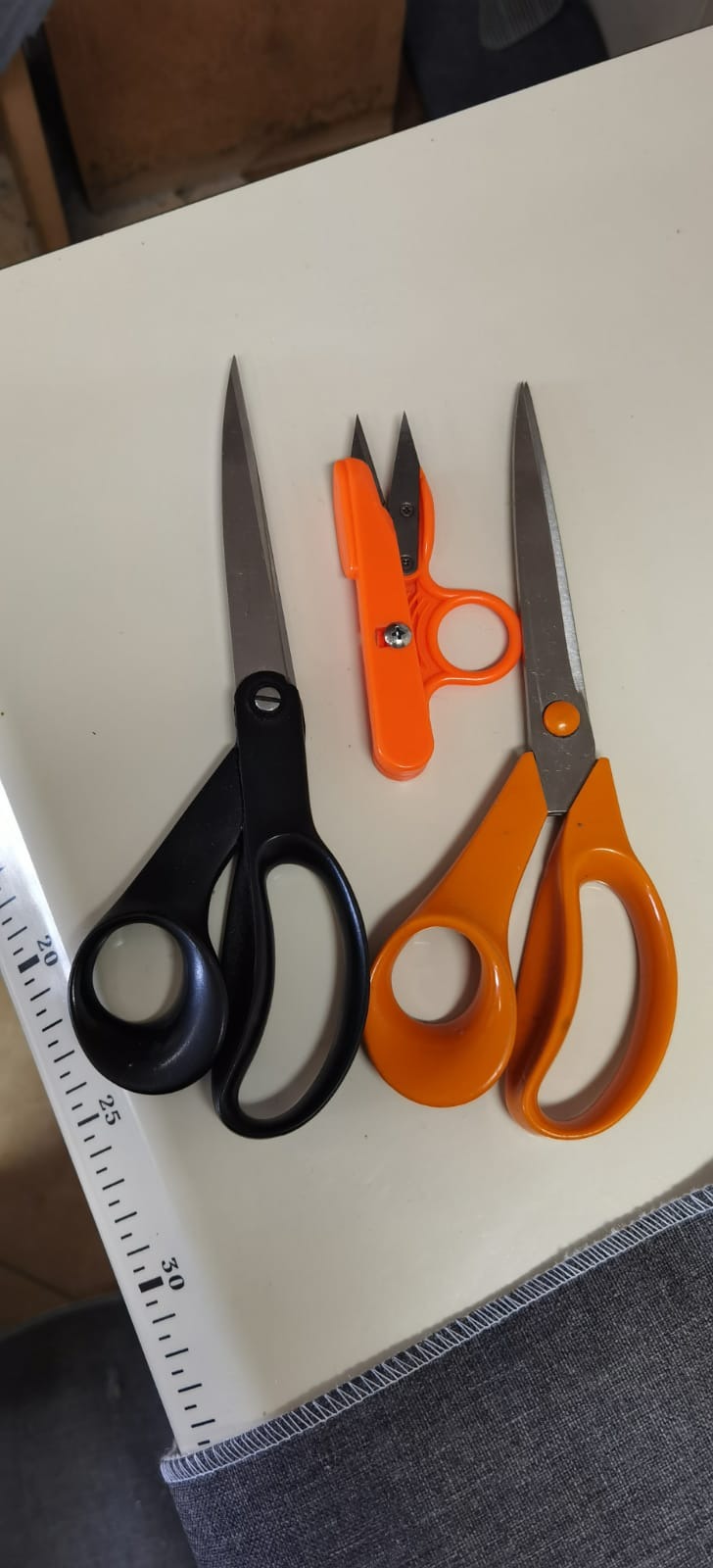 Trzy rodzaje nożyczek krawieckich: czarne, pomarańczowe i małe pomarańczowe do prucia szwów, leżące na stole obok fragmentu szarego materiału z widocznym szwem brzegowym i linijki.