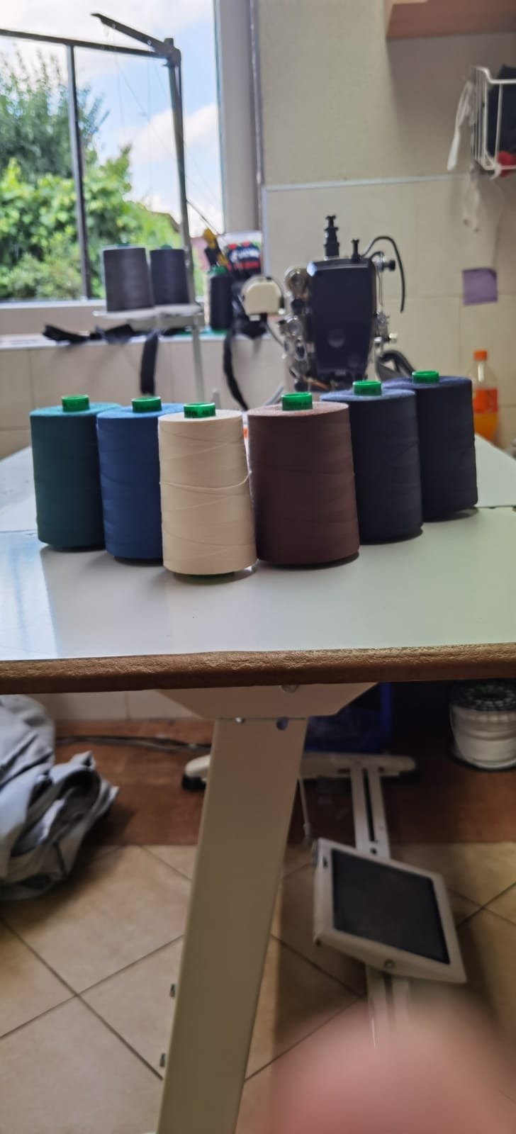 Rząd kolorowych nici na szpulach, ustawionych na białym stole krawieckim, z maszyną do szycia w tle i widokiem z okna na zieleń.