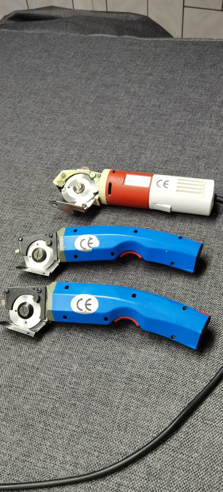 Trzy elektryczne noże tarczowe do cięcia tkanin, jeden z czerwono-białą obudową, dwa z niebieską, leżące na szarej tkaninie.