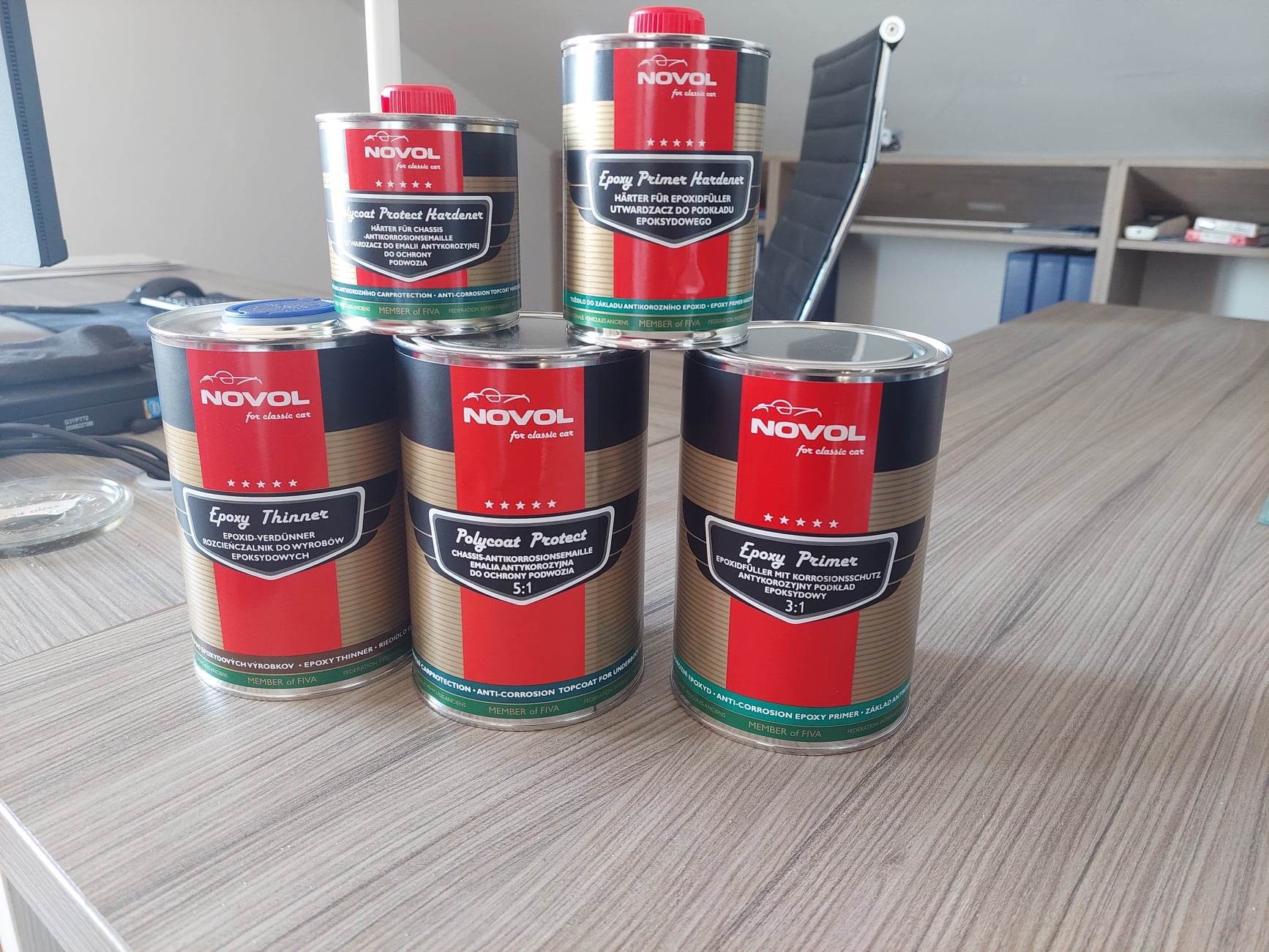 Pięć puszek z preparatami marki Novol dla klasycznych samochodów: Epoxy Thinner, Polycoat Protect i Epoxy Primer, ustawione na drewnianym biurku w biurze.