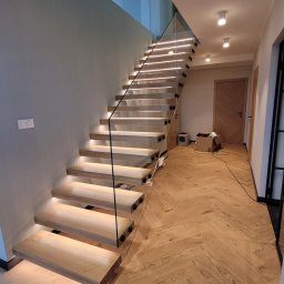 PREFABRICADO - Nowoczesne schody drewniane z podświetleniem LED i szklaną balustradą, widok z dołu na górę, parkiet w jodełkę na podłodze, elementy montażowe balustrady w kolorze czarnym.