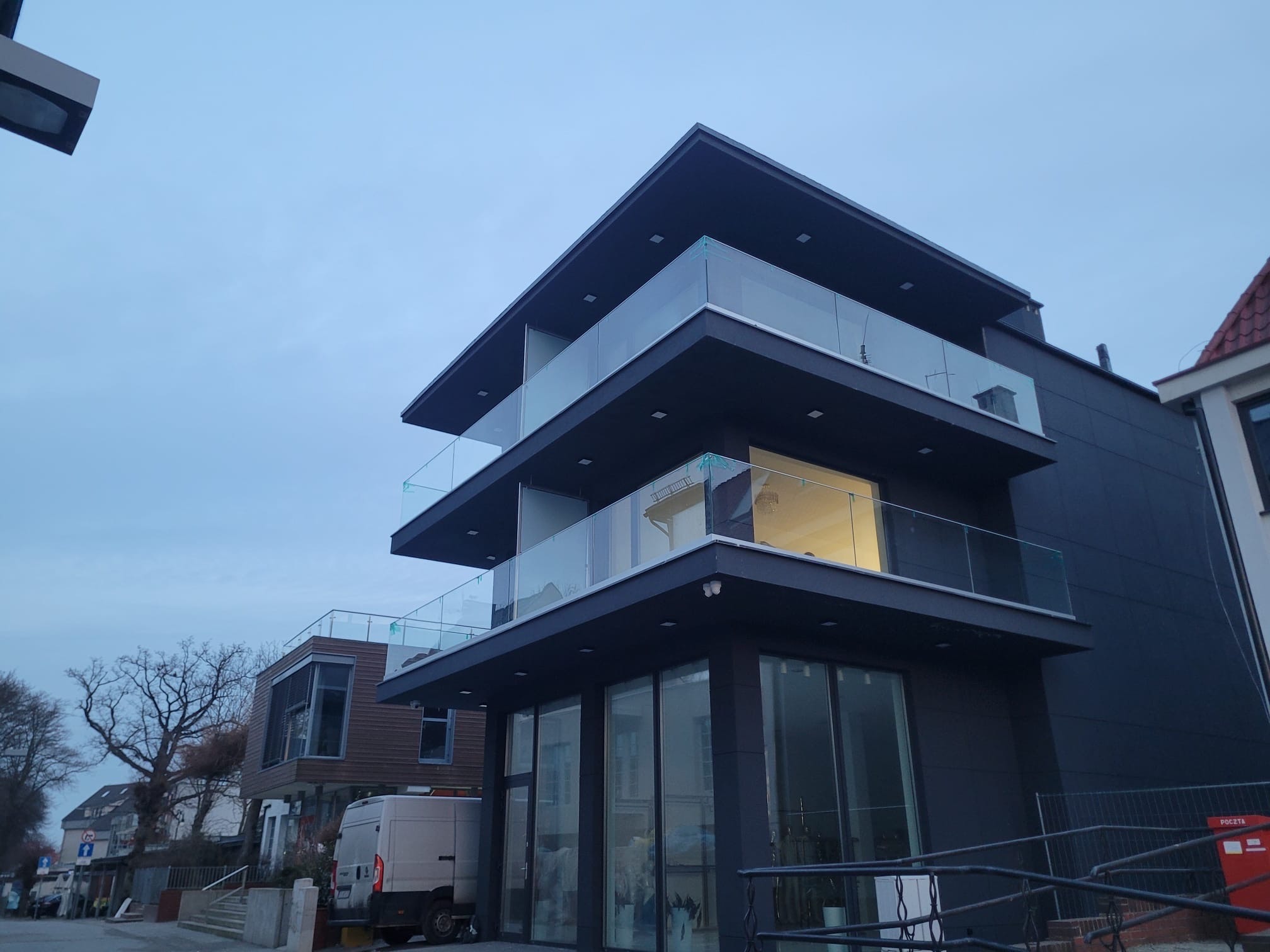 Nowoczesny budynek z trzema kondygnacjami balkonów zabezpieczonych przezroczystymi taflami szkła, sfotografowany o zmierzchu.