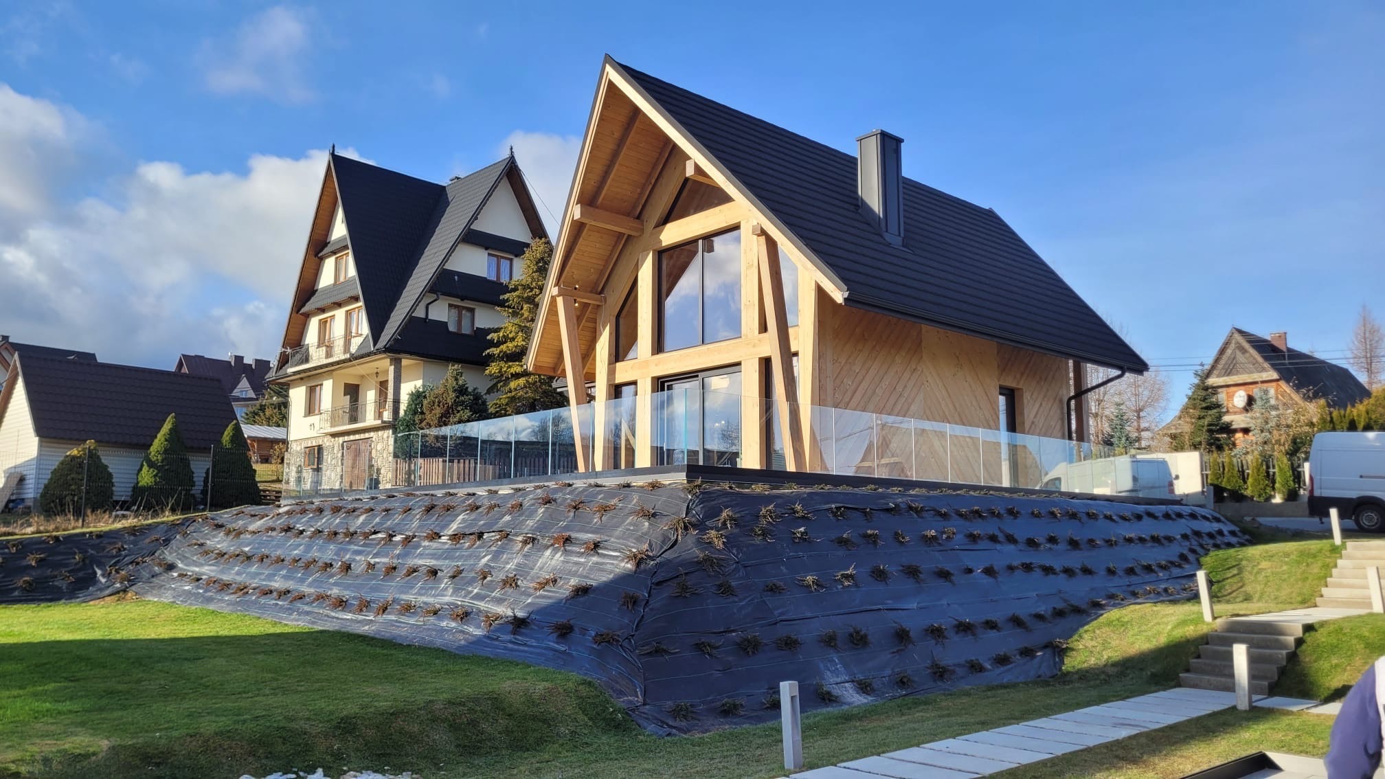 Nowoczesny dom z drewnianą elewacją i szklaną balustradą tarasu, widok z poziomu ogrodu z zabezpieczeniem nasadzeń na skarpie.