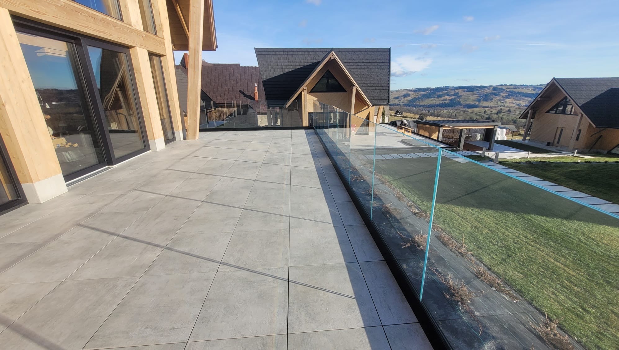 Szklana balustrada na tarasie nowoczesnego domu z widokiem na góry, z elementami drewnianymi i szarą płytką podłogową.