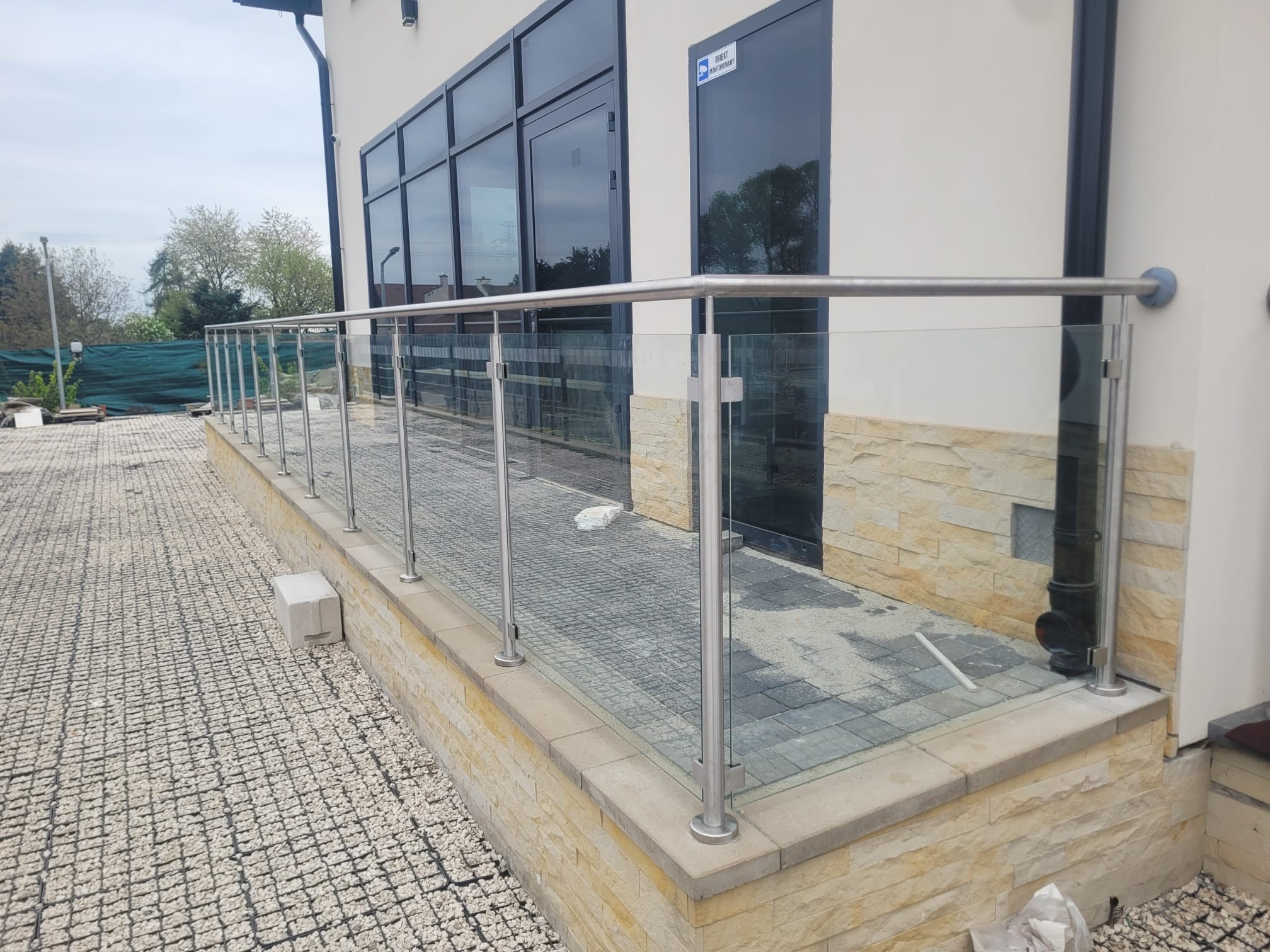 Szklana balustrada ze stalowymi słupkami na tarasie z kostki, widok na budynek z dużymi oknami.