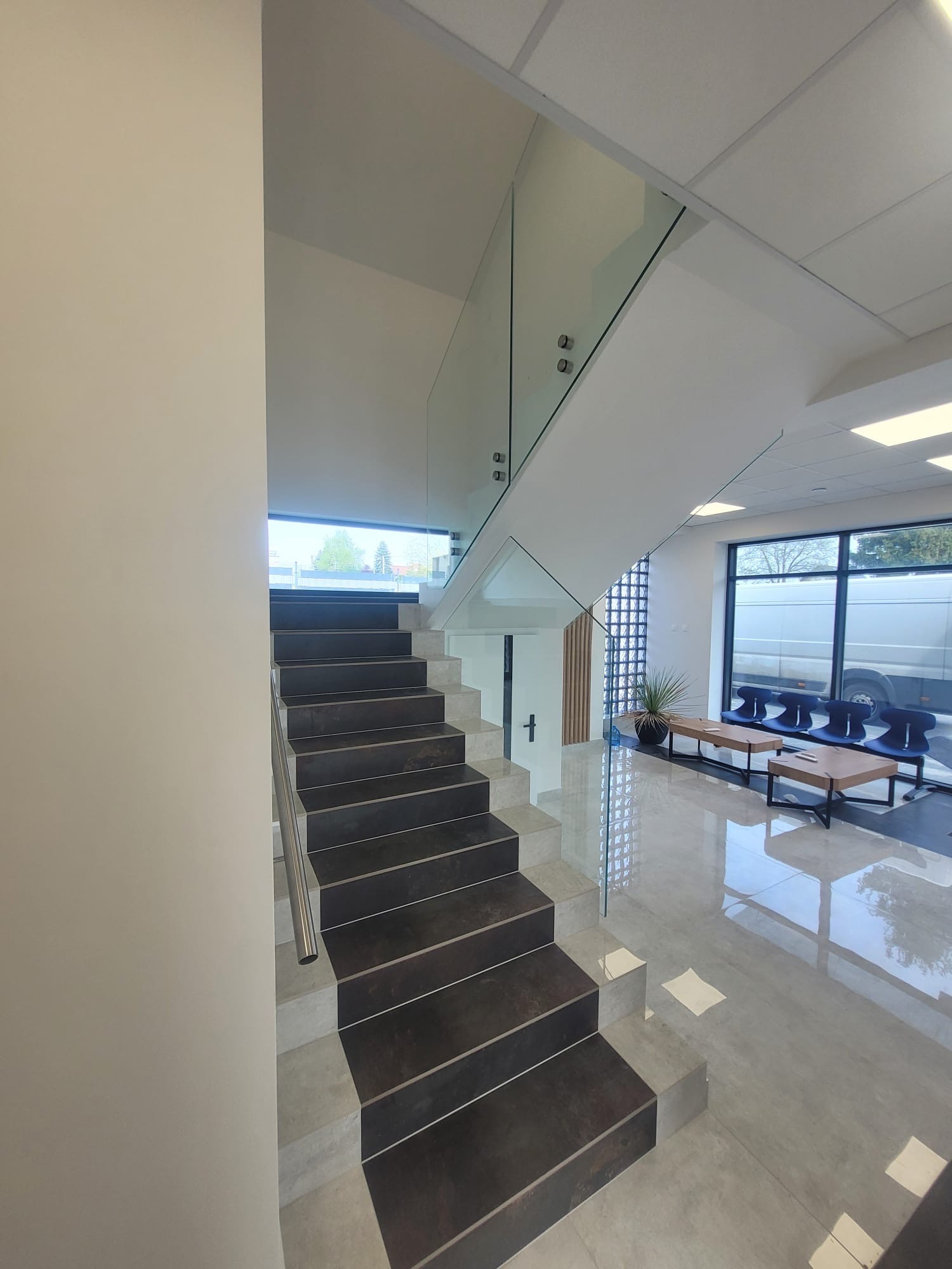 Nowoczesne schody z ciemnymi stopniami, jasnymi podstopnicami, szklaną balustradą mocowaną punktowo i metalową poręczą w przestronnym, jasnym holu biurowym.