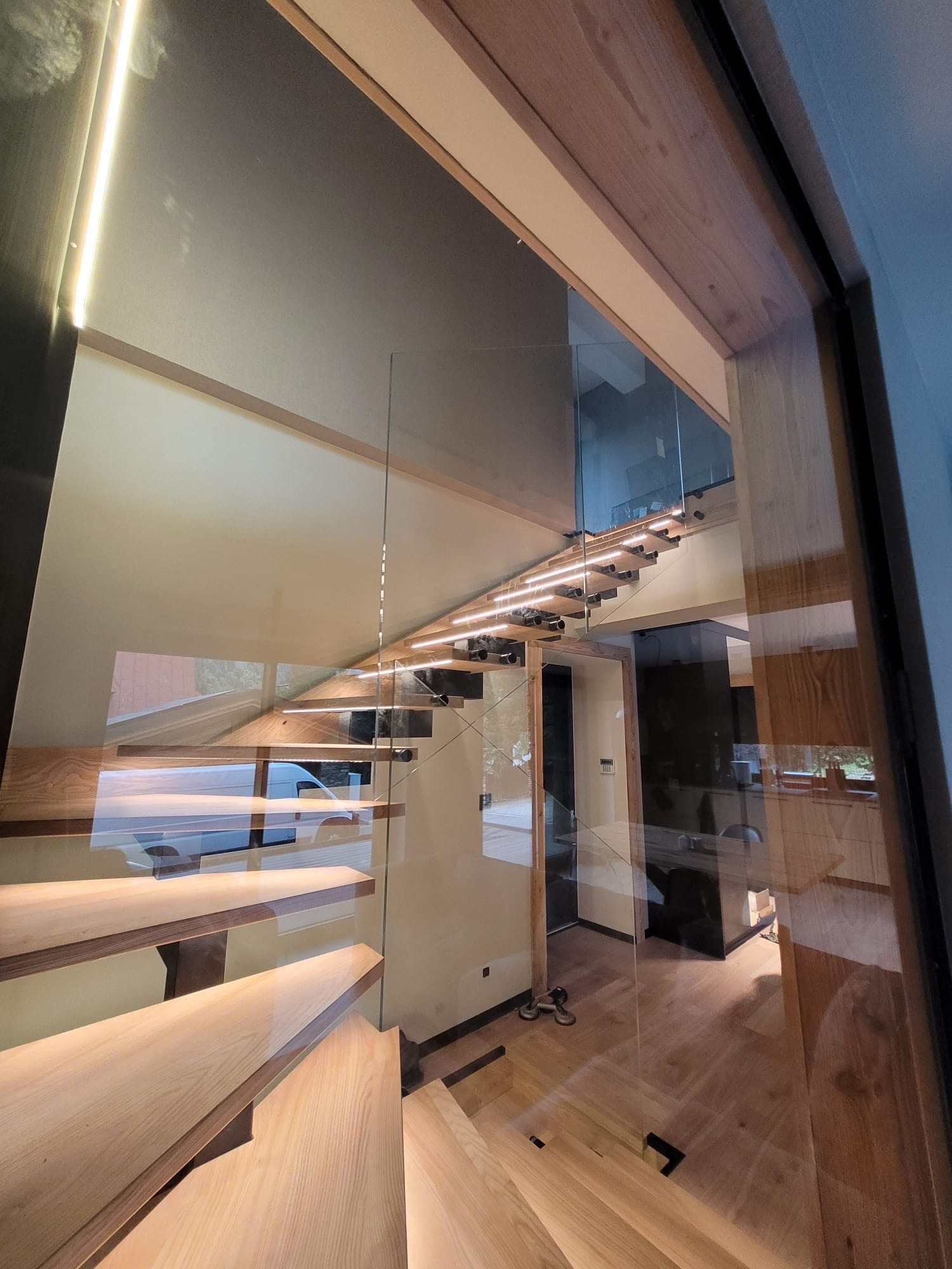 Nowoczesne schody drewniane z podświetleniem LED, zabezpieczone szklaną balustradą, widoczne z perspektywy dolnej części biegu.