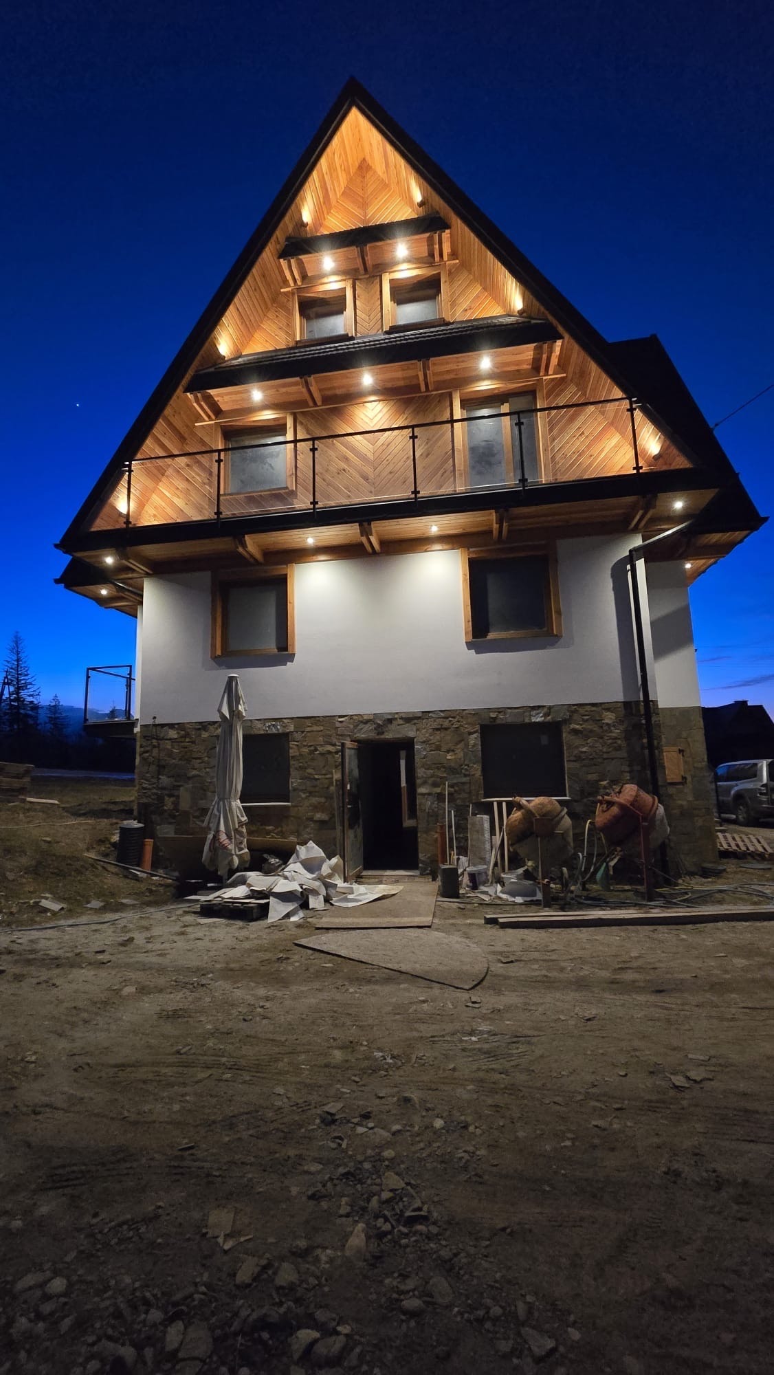 Nocny widok na dwupiętrowy dom z drewnianym wykończeniem i balkonem zabezpieczonym czarną balustradą, częściowo wykończony kamieniem, z widocznymi śladami prac budowlanych.