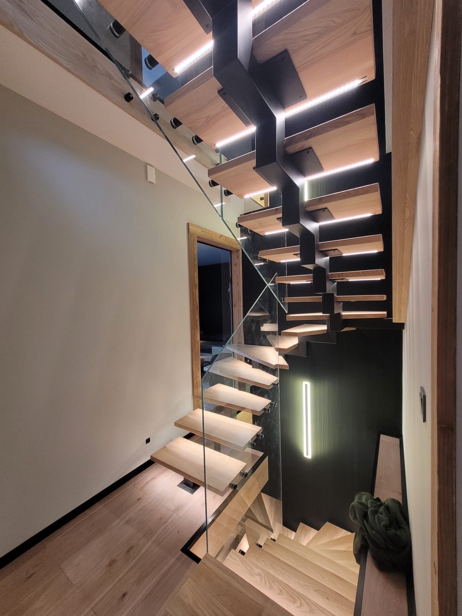 Nowoczesne schody drewniane z podświetleniem LED i szklaną balustradą, widok z dołu, minimalistyczne wnętrze z ciemną ścianą akcentującą.