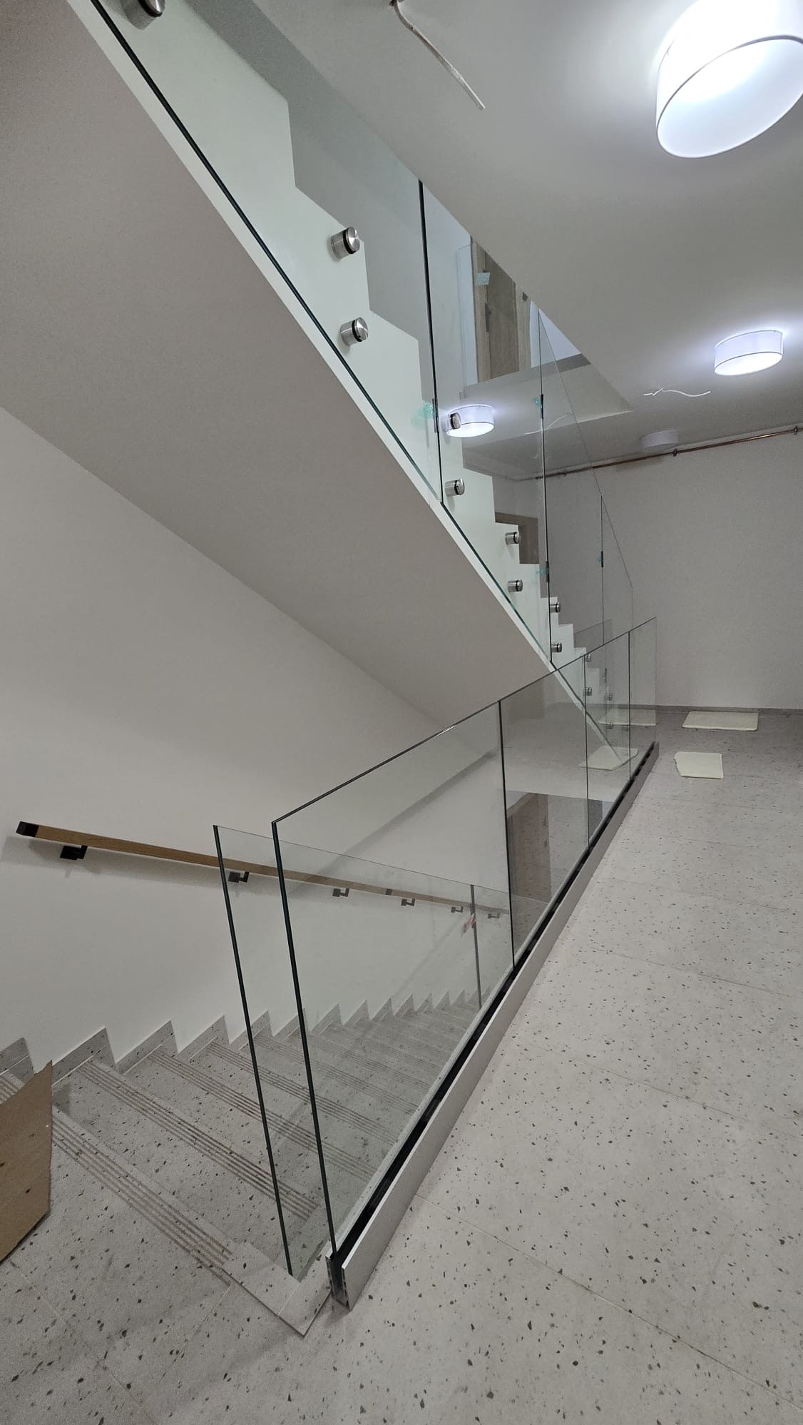 Szklana balustrada schodowa z drewnianą poręczą, mocowana punktowo, widok z dołu na jasnym, terazzo podłożu.