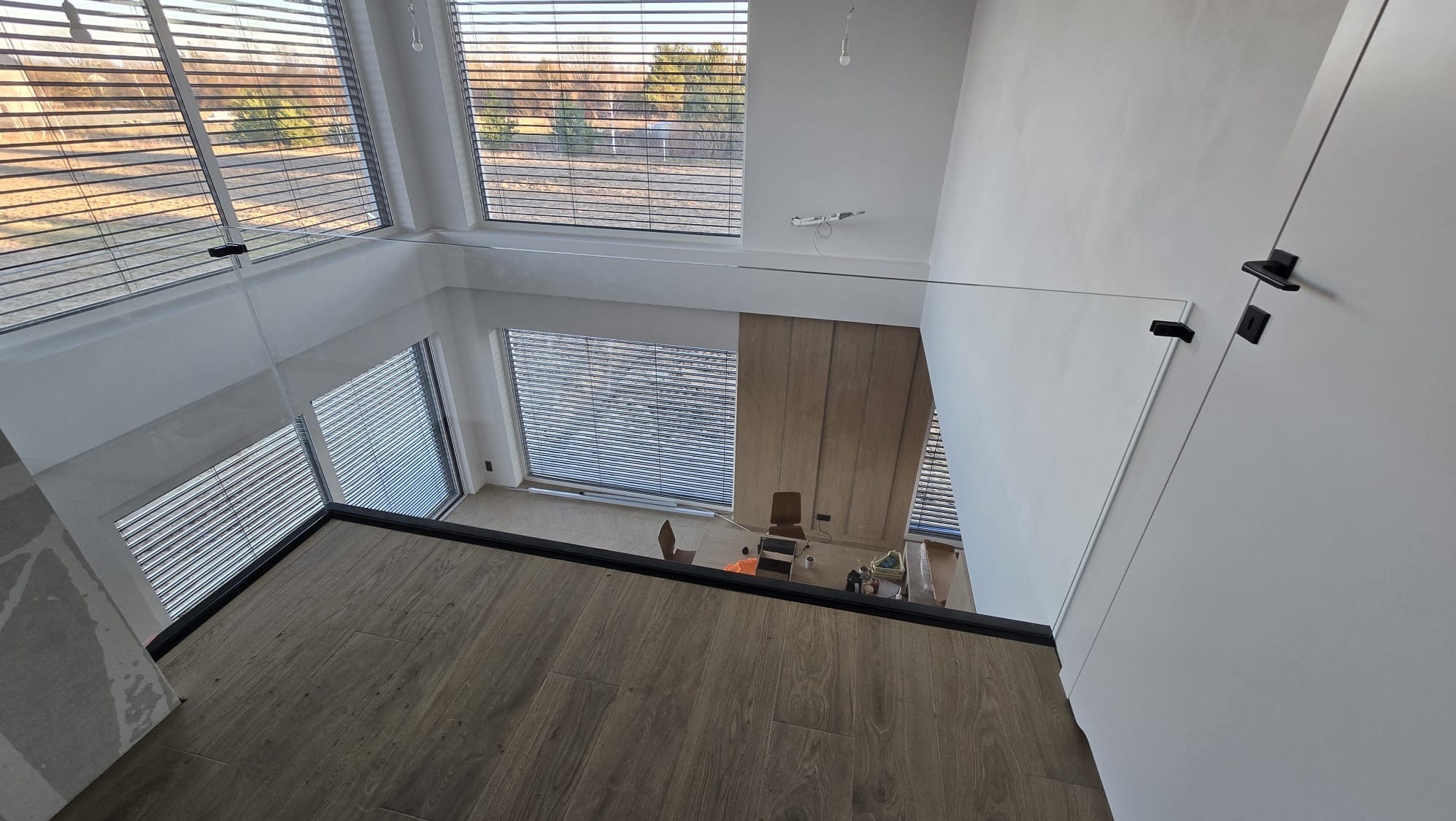 Wysoki kadr na szklaną balustradę w nowoczesnym wnętrzu z dużymi oknami i drewnianą podłogą, widok z góry na niższy poziom z biurkiem i krzesłem.