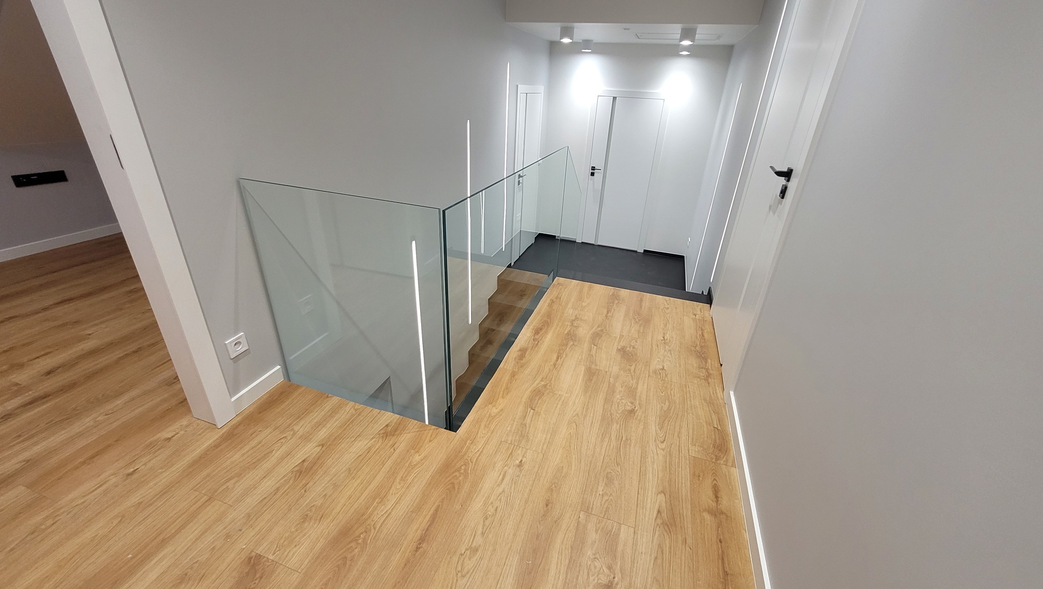 Nowoczesne schody z drewnianymi stopniami i szklaną balustradą z wbudowanym oświetleniem LED, widok z góry na jasne wnętrze z białymi ścianami i drzwiami.