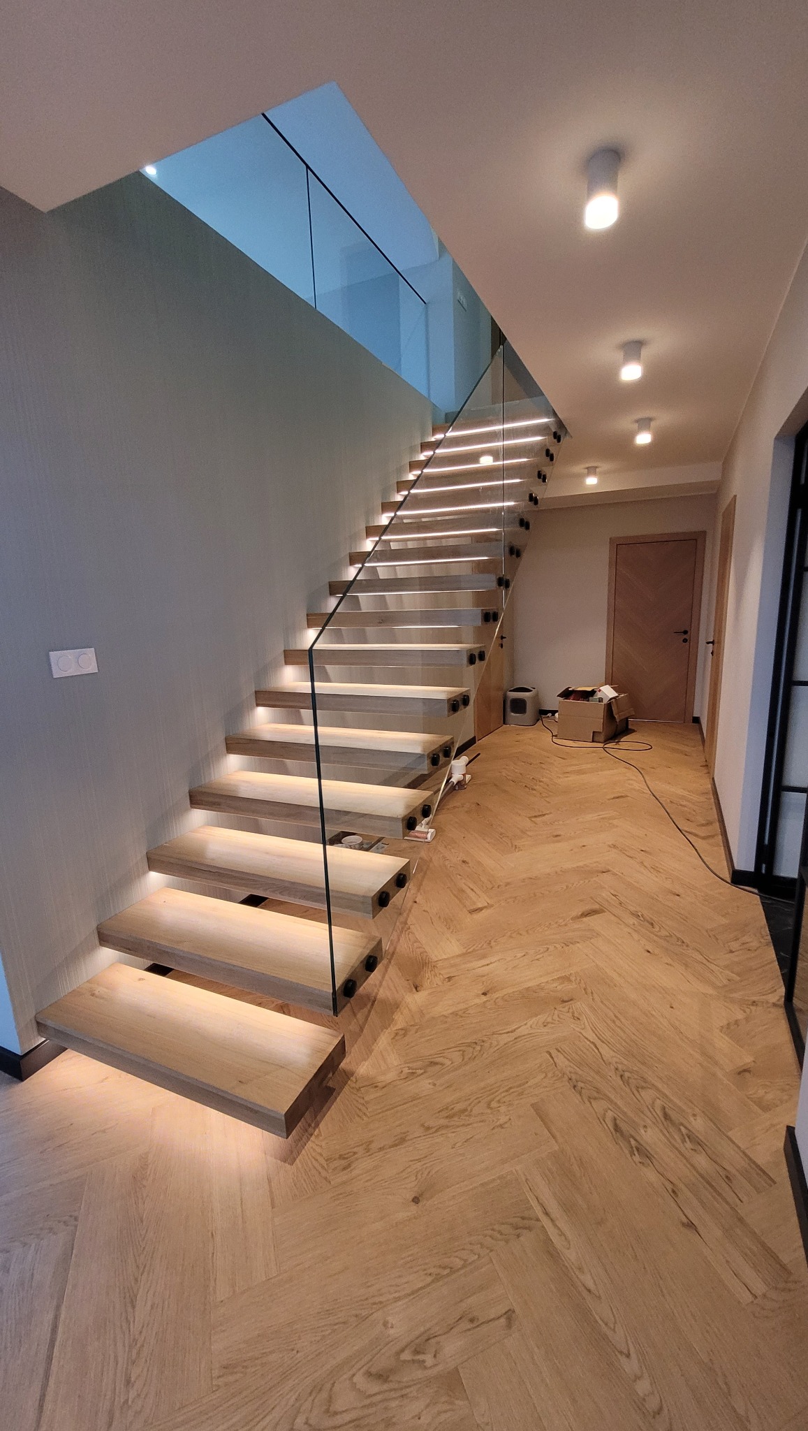 Nowoczesne schody drewniane z podświetleniem LED i szklaną balustradą, widok z dołu na górę, parkiet w jodełkę na podłodze, elementy montażowe balustrady w kolorze czarnym.