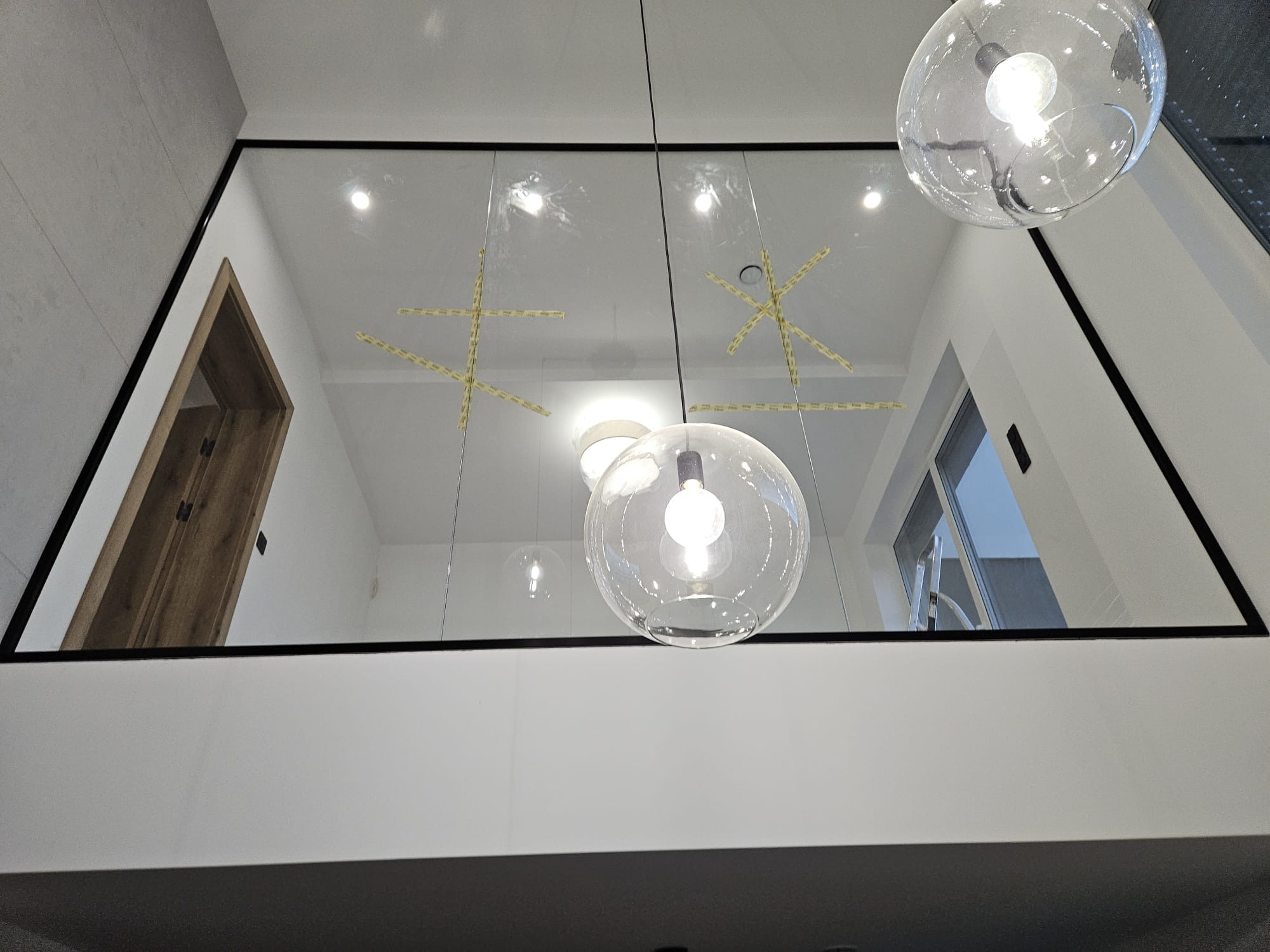 Nowoczesna szklana balustrada z czarną ramą, widok z dołu, z wiszącymi szklanymi lampami i widocznymi taśmami zabezpieczającymi na szkle.