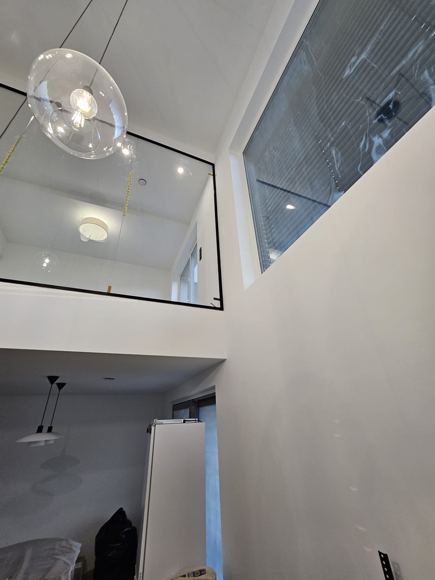 Wnętrze domu z widoczną szklaną balustradą na antresoli, oświetloną lampą wiszącą w kształcie kuli i oknem z żaluzjami.